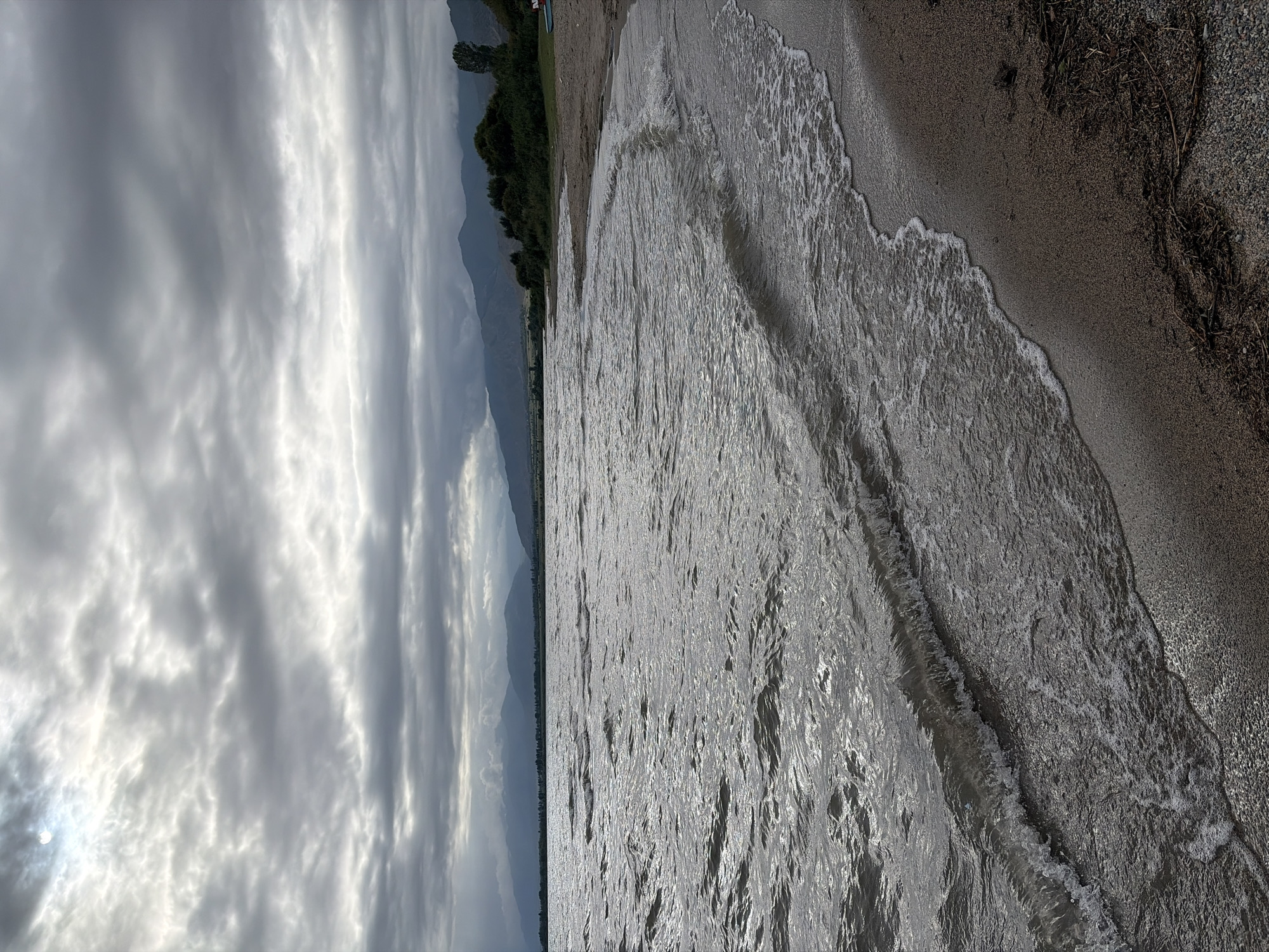 An overcast sky with dramatic, textured clouds looms over a tranquil lake with choppy water. The sun is partially visible, creating a bright spot through the clouds on the upper left side. In the distance, rolling hills and mountains are shrouded in haze. A narrow, sandy beach with scattered debris runs along the right side of the frame, meeting the water's edge. The overall mood is somber and atmospheric.