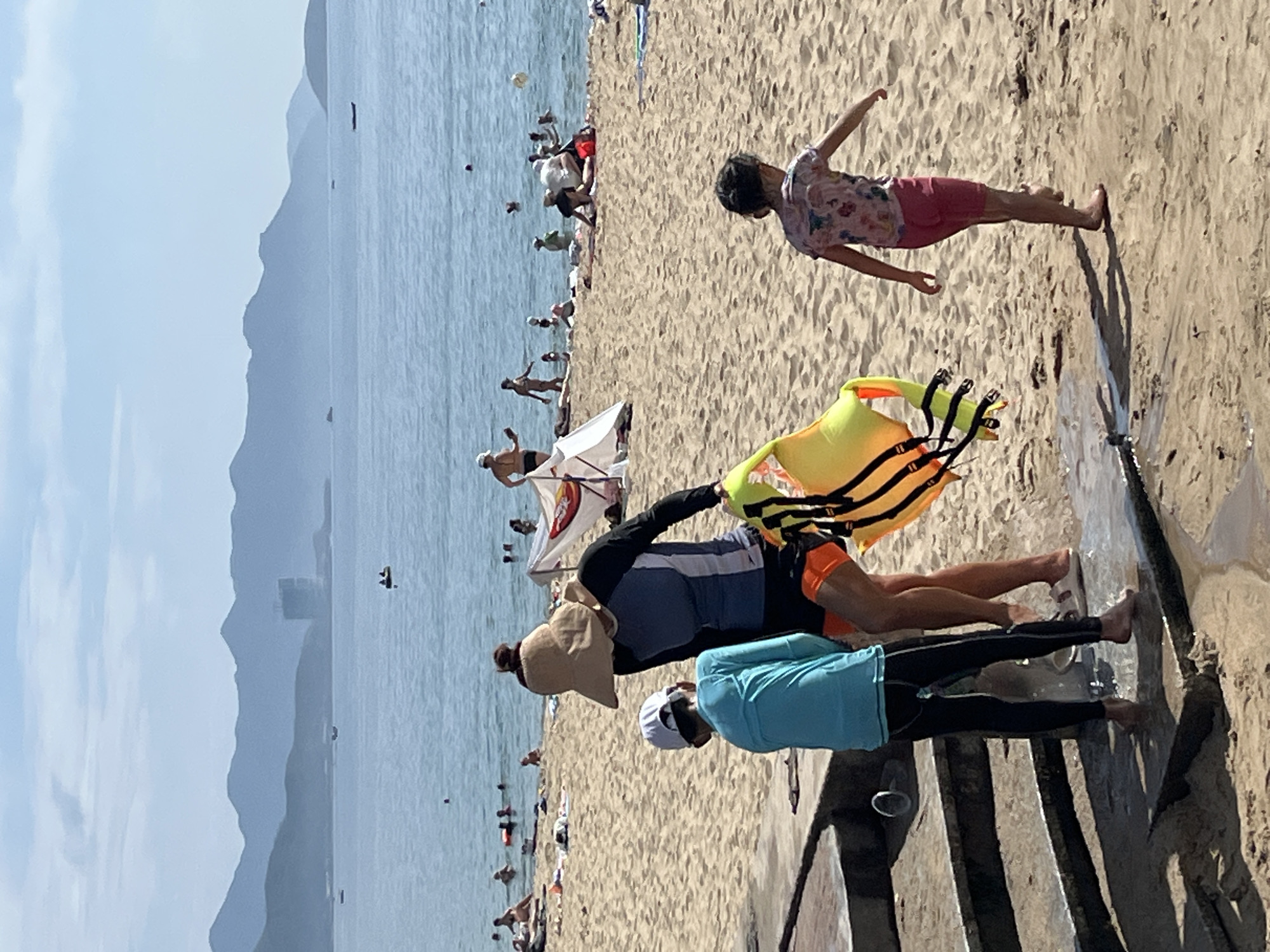 The image depicts a sunny day at a beach in Nha Trang, Vietnam. In the foreground, a person wearing a blue long-sleeved shirt and black pants stands on a wet concrete surface. To their right, another person in a dark blue rash guard, orange shorts, and a wide-brimmed hat holds a yellow and orange life vest. They are both facing away from the camera, towards the water. A young girl in a colorful patterned shirt and pink shorts walks away from them on the sand. The beach is populated with numerous other people, some swimming in the clear blue ocean, others relaxing on towels or under umbrellas. In the background, gentle waves lap against the shore, and a line of hazy, green mountains stretches across the horizon under a pale blue sky. Boats are visible on the water, and some structures are present on the distant coastline. The overall scene is one of leisure and recreation on a tropical beach.
