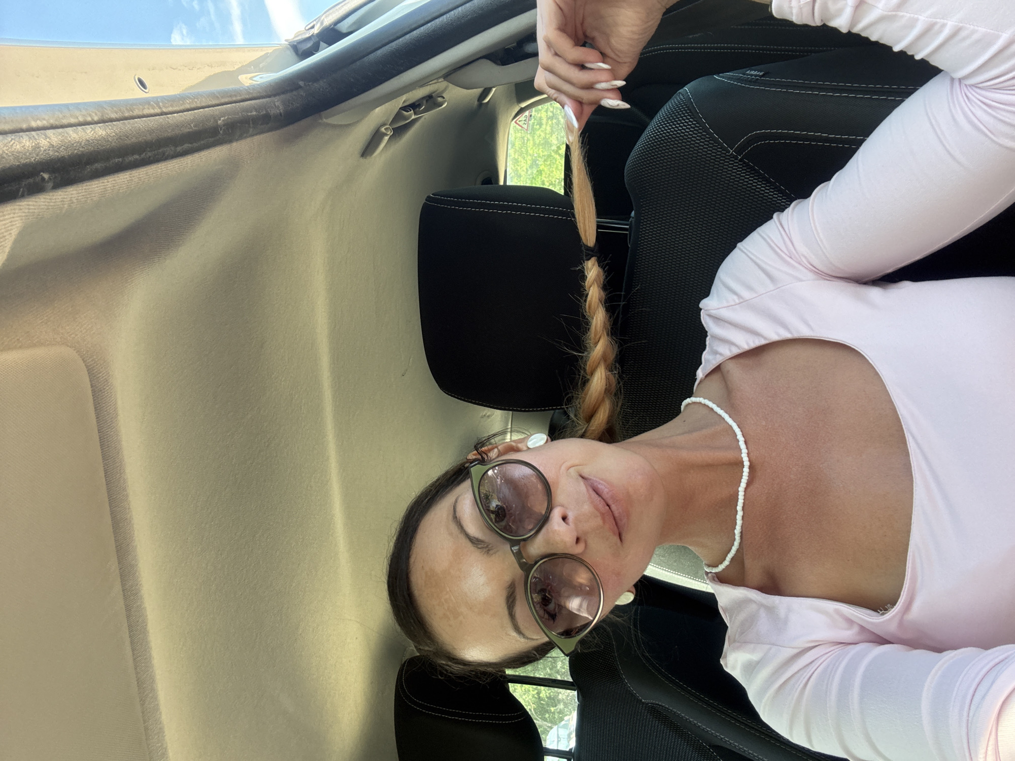 A woman with brown hair is sitting in the driver's seat of a car. She is wearing sunglasses, a pink top, and a white beaded necklace. Her hair is in a braid that she is holding with her right hand. The car's interior is visible, including the seats and the headliner. The exterior of the car is also visible through the windows, showing trees and a blue sky. The image appears to have been taken during the day.