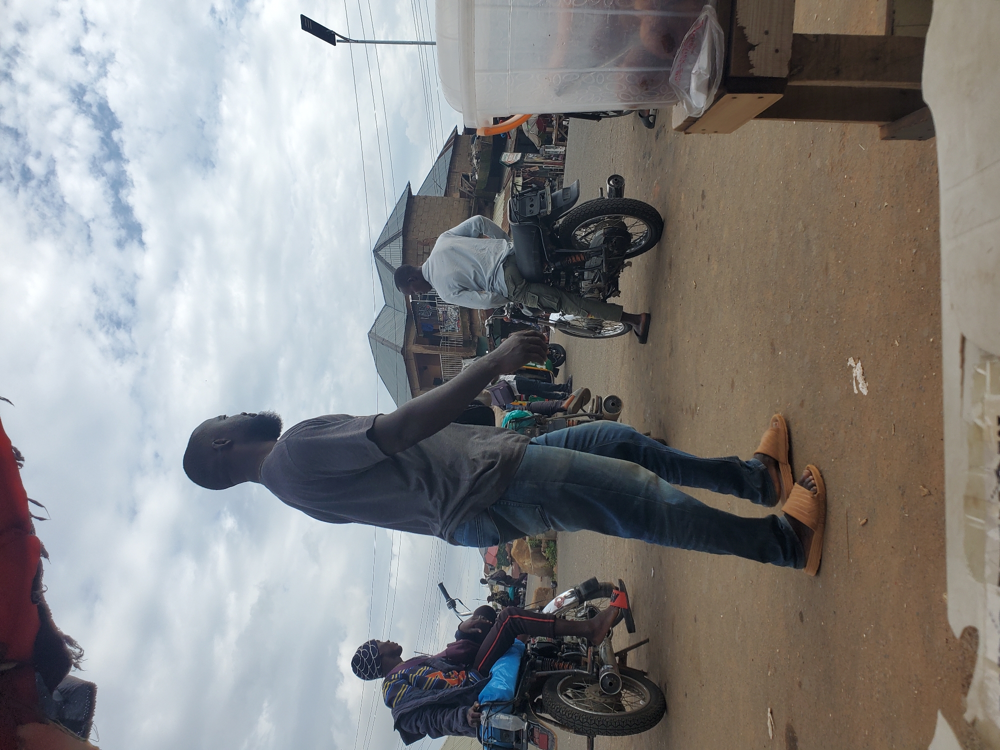 A man is walking on a dirt road in Gwong, Nigeria. He is wearing a gray t-shirt, blue jeans, and brown sandals. He has a beard and short hair. In the background, other people are riding motorcycles and standing around. Buildings with corrugated metal roofs are visible in the distance. The sky is cloudy.