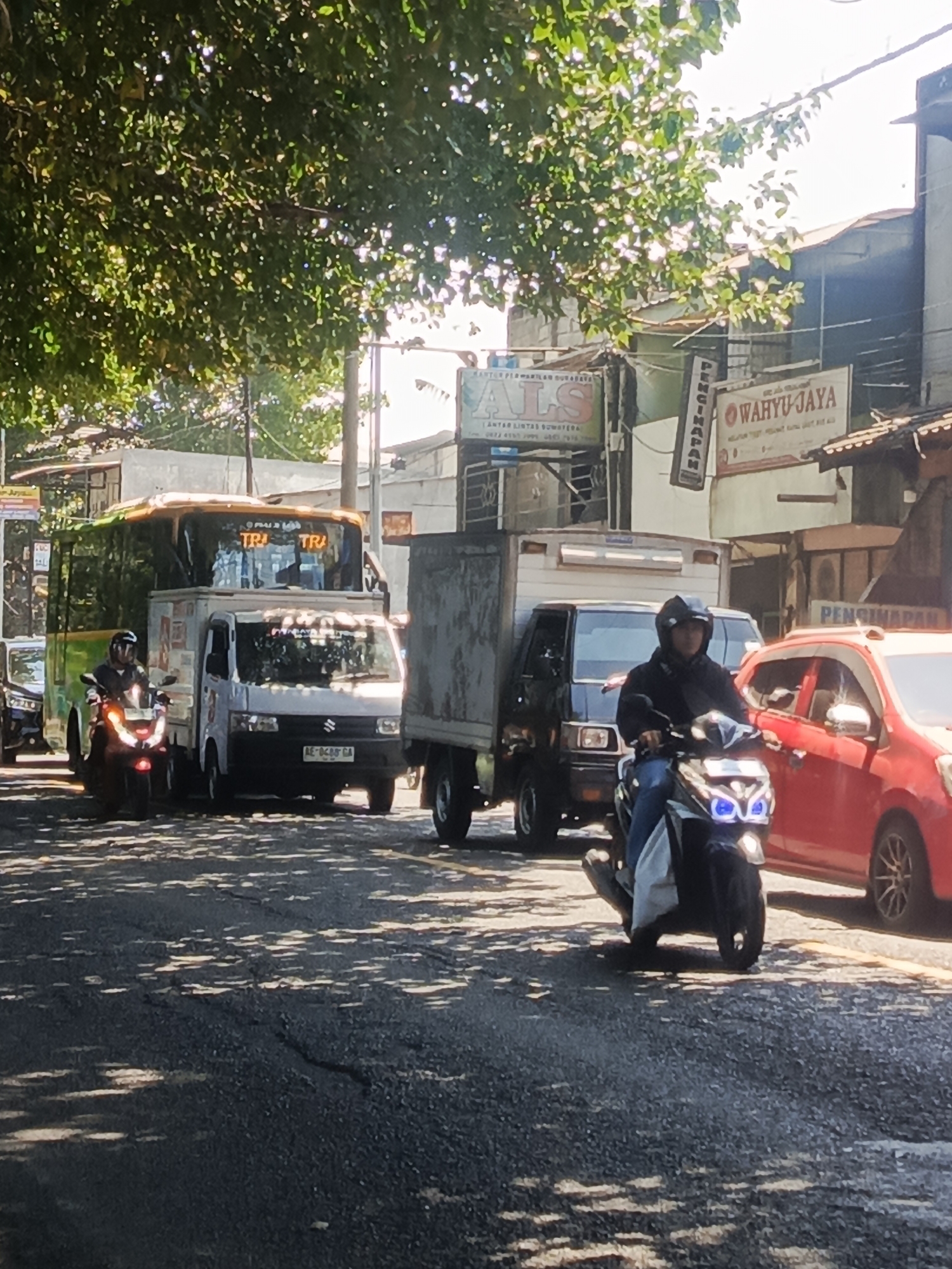 The image depicts a traffic scene in Bungurasih, Indonesia, likely near the Bungurasih Bus Terminal, a major transportation hub. The foreground shows a dark asphalt road with patches of sunlight filtering through a leafy tree canopy, creating dappled shadows on the surface. A motorcycle rider, wearing a dark jacket and helmet, is positioned in the lower right of the frame, the front of their scooter illuminated by bright blue headlights.
In the midground, several vehicles are visible in traffic. A white truck with a grey cargo box is positioned directly behind the motorcycle. To the left of this truck, a bus with an orange top and green lower section is partially obscured, displaying "TRI TRA" on its front. Further left, another vehicle, possibly a car, is visible.
The background reveals a bustling urban environment with buildings and signage. One sign prominently displays "WAHYU JAYA" in red and white lettering. Another sign, partially visible, has the text "ALS" and "ANTAR LINTAS SUMATERA". Structures appear to be a mix of concrete and corrugated metal, consistent with a commercial or industrial area. The overall atmosphere suggests a busy daytime traffic situation.