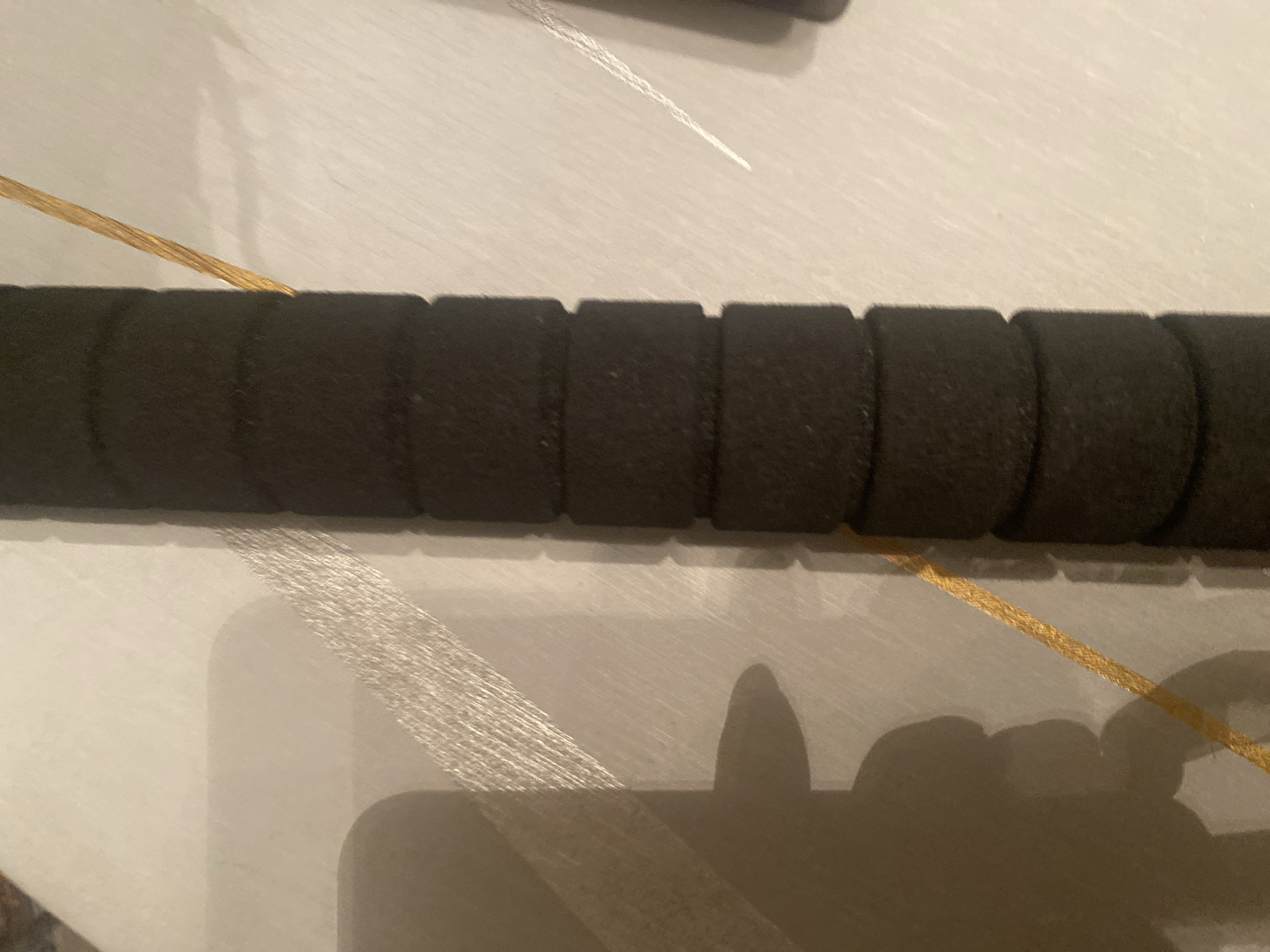 The image shows a black foam grip with segmented sections, likely from a tool or exercise equipment, lying on a light-colored textured surface. A thin, golden braided cord runs diagonally across the frame, passing behind the foam grip. To the left of the grip, a shadow of a hand or arm is cast on the surface. The scene is illuminated by a light source from the left, creating the shadow and highlighting the texture of the surface. The background is out of focus. The location in Chernogorsk, Russia is not discernible from the image.