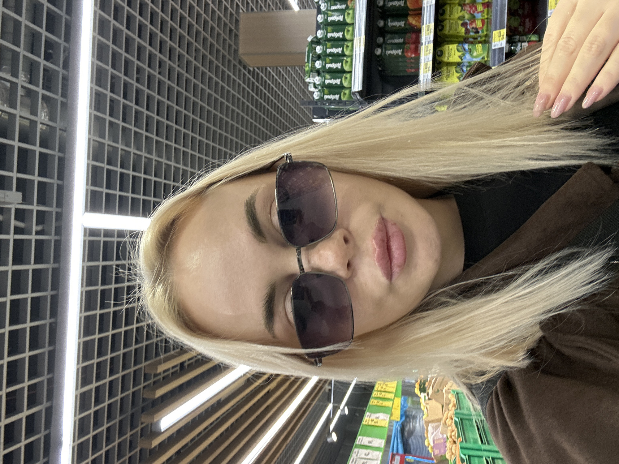 A blonde-haired woman wearing sunglasses is in a store aisle in Megion, Russia. Shelves of products are visible in the background.