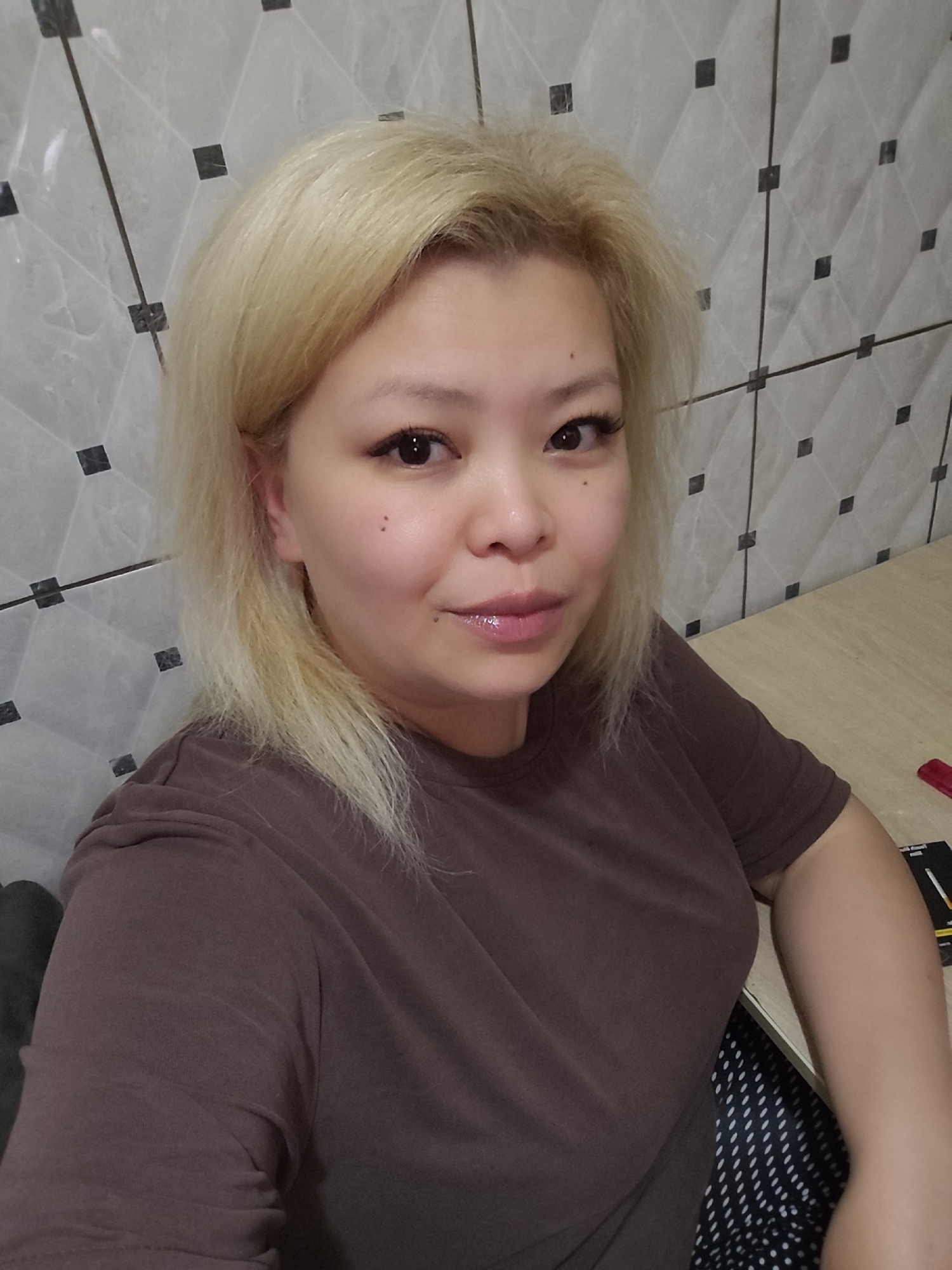 A woman with fair skin and blonde hair is centered in a selfie-style photograph. She wears a brown T-shirt. The background features a tiled wall with a geometric pattern of white tiles with small black square inlays and a portion of a light-colored wooden desk. Other items are visible on the desk but are unidentifiable. The image provides no information to place the location in Sarıyer, Turkey.