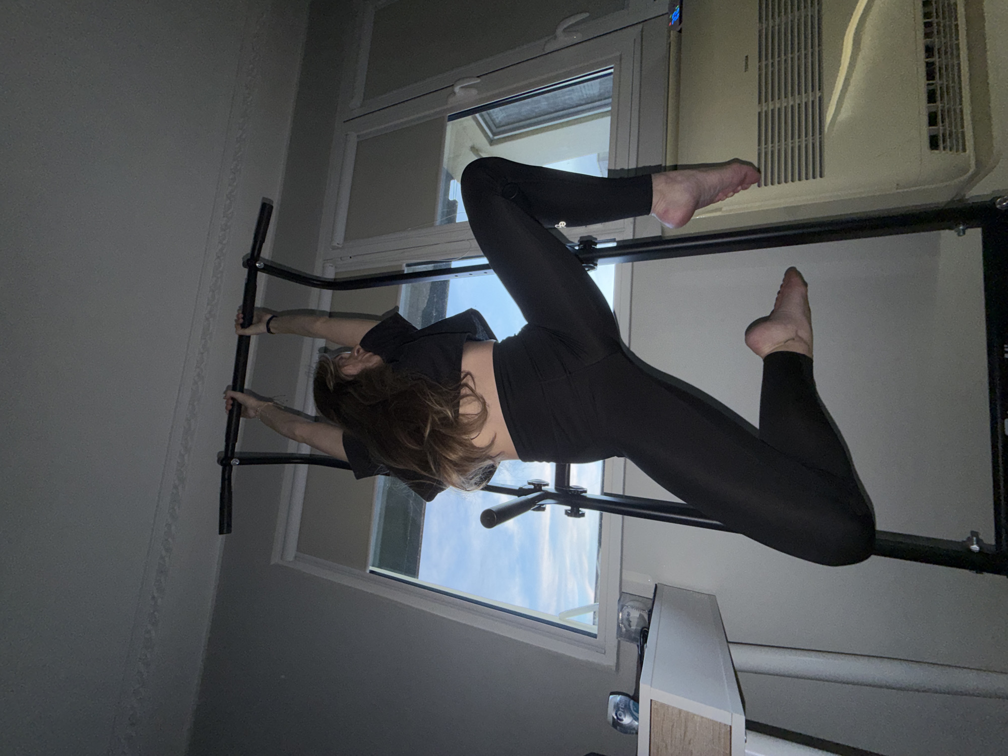 A woman in black leggings and a black top is suspended upside down on a black pull-up bar, with her feet raised towards an air conditioning unit. The bar is positioned within a room featuring a window with a view of a blue sky. The room's interior includes white walls and what appears to be a white cabinet. The location is not identifiable as being in Plovdiv, Bulgaria.