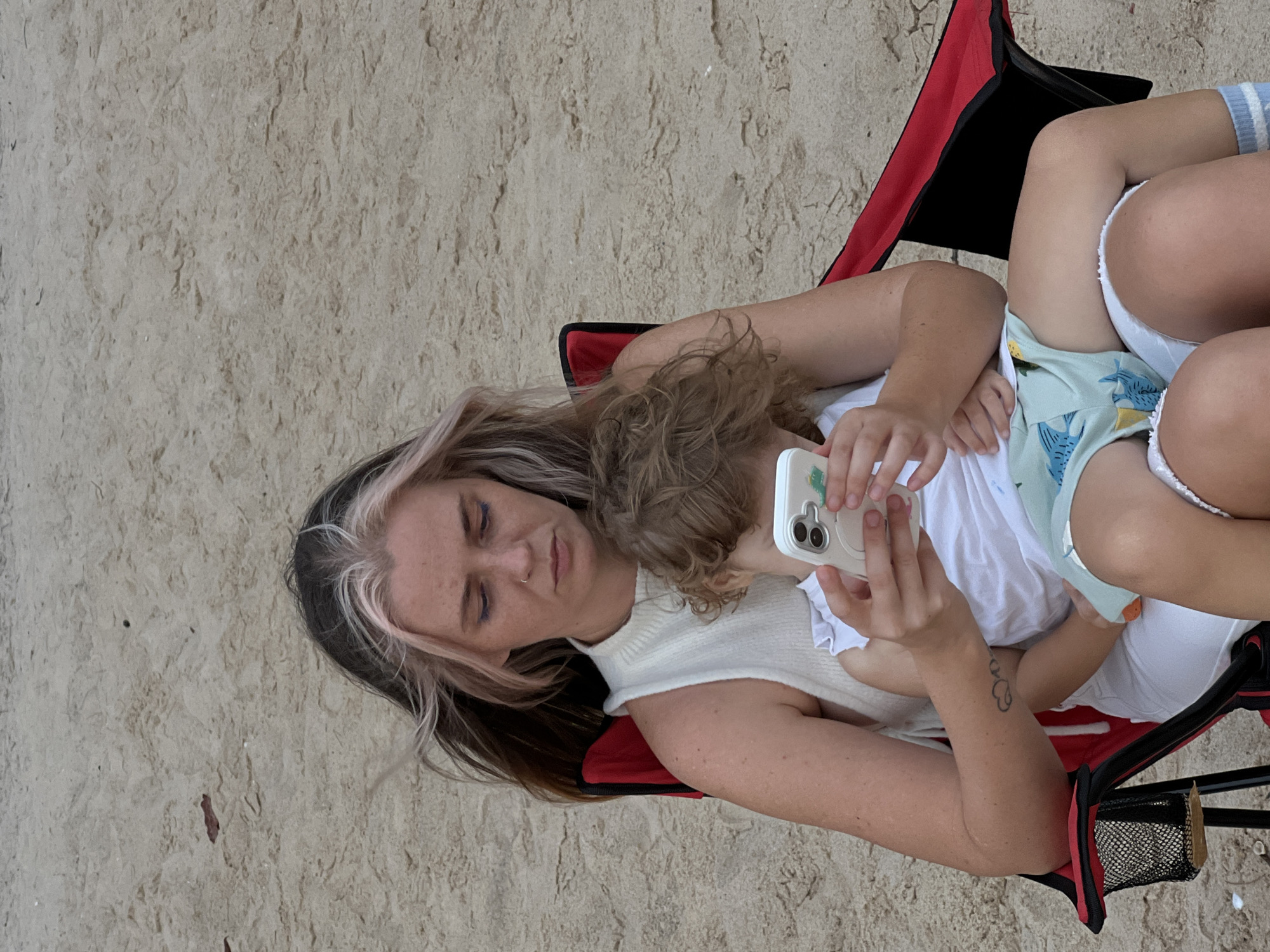 A woman and a child are sitting on a beach in a red camping chair. The woman is holding a smartphone, and the child is looking at the phone. The woman has long, dark hair with blonde highlights and is wearing a white sleeveless top. The child has curly blonde hair and is wearing a white t-shirt and light blue shorts with a shark print. The beach is covered in sand, and the background is out of focus. The image appears to be taken in daylight.