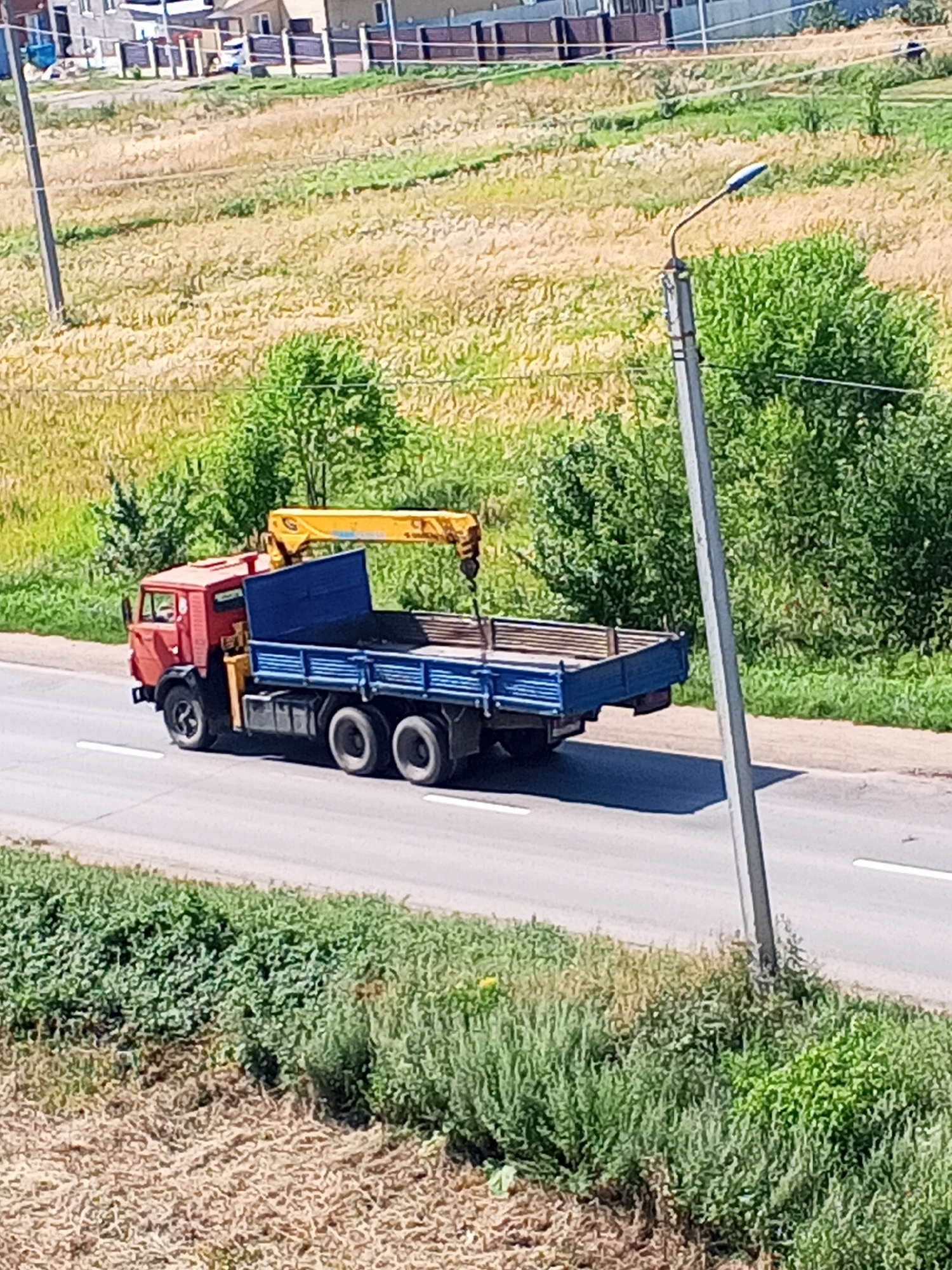 The image shows a red truck with a blue flatbed trailer and a yellow crane attached, driving on a paved road. The truck is positioned in the center of the frame and is moving from left to right. The road has white lane markings. On the left side of the road, there is a grassy embankment with dry straw-like vegetation. On the right side of the road, there is a stretch of green bushes and trees. In the background, there is a field of dry grass and some buildings. A lamppost is visible on the right side of the road, leaning slightly. The scene appears to be in a rural or suburban area, possibly on the outskirts of Chistopol, Russia. The sky is not visible in the frame. There are no people clearly visible in the image.