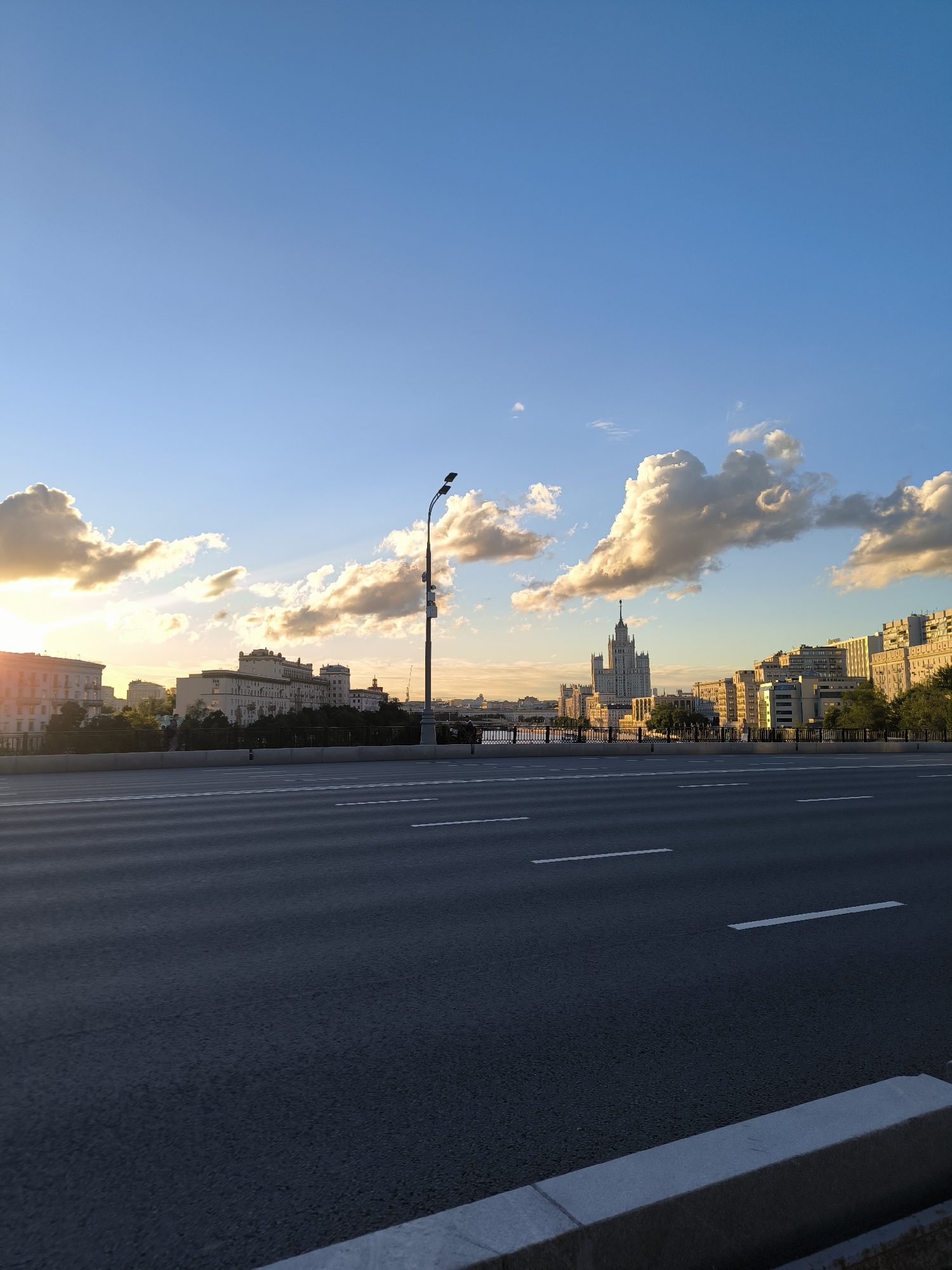 This media file captures an urban landscape in Moscow, Russia, during what appears to be late afternoon or early evening, indicated by the warm, golden light of the setting sun. The sky is a vibrant blue, adorned with dramatic, illuminated clouds that add depth and texture to the scene.
The foreground is dominated by a multi-lane asphalt road, marked with white dashed lines. The road stretches towards the horizon, suggesting a vast cityscape. A concrete curb runs along the edge of the road.
In the midground, a series of buildings line a waterfront, with trees providing a natural buffer. A notable structure in the distance is a tall, distinctive skyscraper with a spire, characteristic of Stalinist architecture, which is a prominent landmark in Moscow. Other buildings of varying heights and architectural styles are also visible, some with warm lighting suggesting that many windows are illuminated.
A lamppost stands on the right side of the road, adding to the urban feel. There are hints of people walking along the waterfront, appearing as small silhouettes against the backdrop of the city. The overall mood is serene and picturesque, with the warm light creating a sense of tranquility and beauty over the bustling city. There is no visible text in the image.