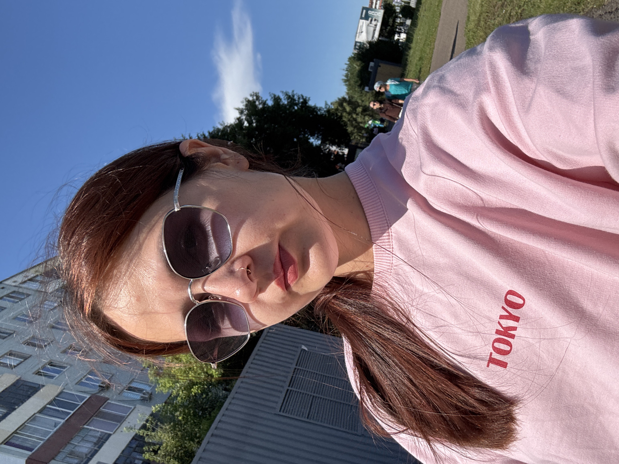 A young woman with reddish-brown hair and sunglasses takes a selfie outdoors on a sunny day in Naberezhnye Chelny, Russia. She is wearing a light pink sweatshirt with the word "TOKYO" in red lettering. Behind her, a multi-story apartment building is visible, along with a gray corrugated fence and some trees. In the background, two other people are walking on a paved path. The sky is a clear blue with a few scattered clouds.
