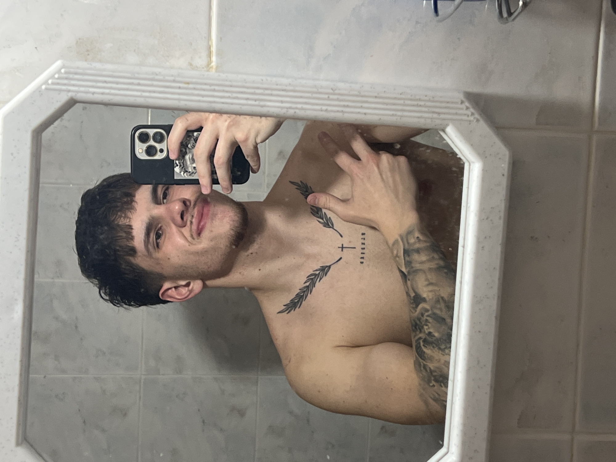 A man is visible in a bathroom mirror. The man is fair-skinned with dark hair and a short haircut. He is shirtless, revealing tattoos on his chest and right arm. The chest tattoo features two olive branches with the word "blessed" printed vertically between them and a cross above. His right arm is covered in a full sleeve tattoo. He is holding an iPhone in his left hand, taking a selfie in the mirror. The bathroom features light-colored tiled walls. The location cannot be determined from this image.