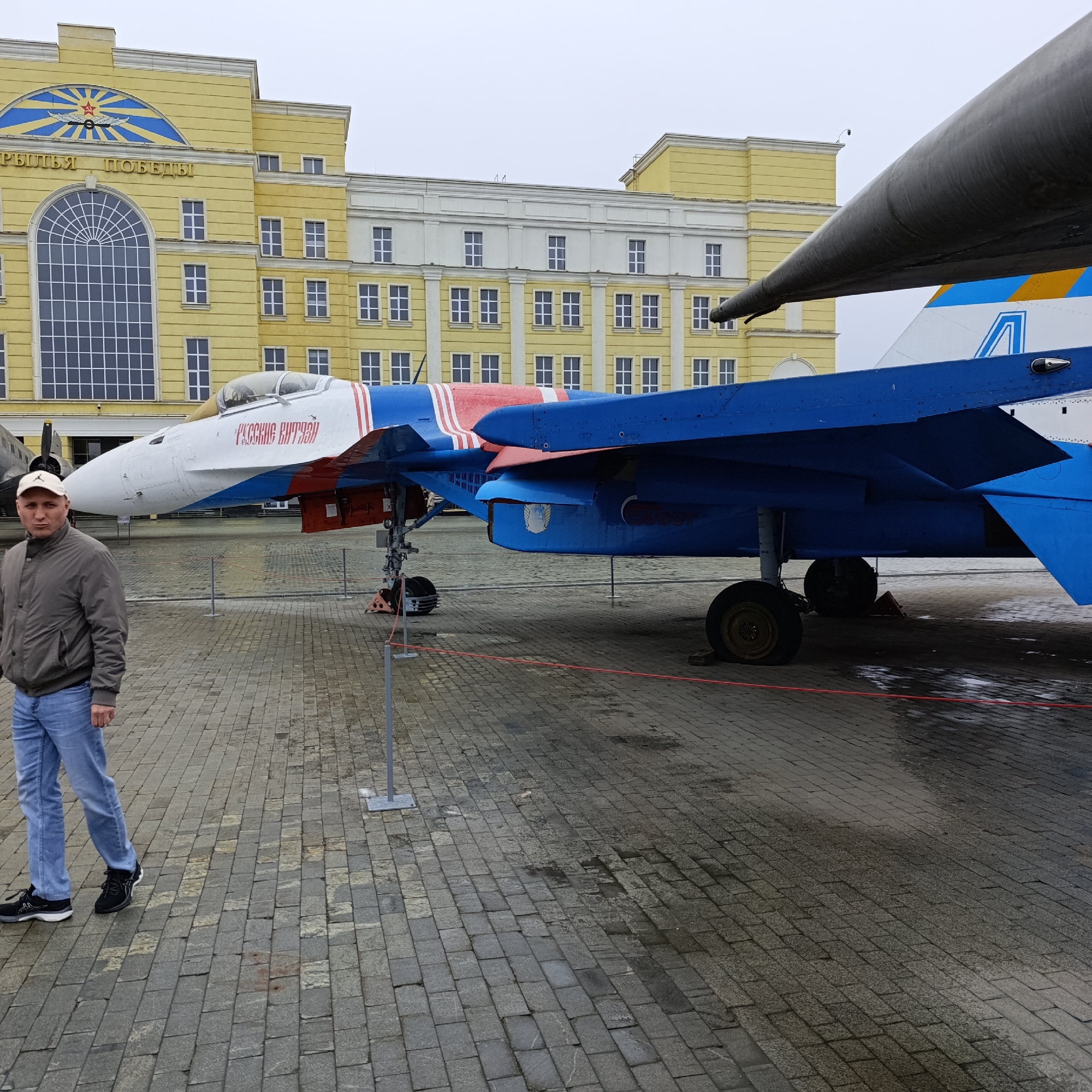 An outdoor, eye-level shot captures a man standing beside a large, blue and white fighter jet, with a yellow and white building forming the backdrop under an overcast sky.
The man, positioned on the left side of the frame, is wearing a light brown baseball cap, a grey jacket, and blue jeans, with black sneakers. He is walking towards the viewer, looking slightly to his left, with a neutral expression. The ground is paved with rectangular stone blocks, appearing somewhat wet in places, suggesting recent rain or damp conditions. A red rope or line runs along the ground, acting as a barrier around the display.
The prominent fighter jet, identified as a Sukhoi Su-27, is painted in a distinctive livery of blue, white, and red stripes, indicative of the "Russian Knights" (Русские Витязи) aerobatic team. The Russian text "РУССКИЕ ВИТЯЗИ" is clearly visible in red on the white section of the aircraft's fuselage, just below the cockpit. The aircraft's landing gear is deployed, and parts of its underside, including a large air intake and additional smaller markings, are visible.
In the background stands a large, multi-story building, primarily painted yellow with white architectural trim. A significant feature of the building is a large, arched window dominating the lower-left section of its facade. Above this window, a semicircular blue emblem with a stylized red star and wings is visible, alongside the golden Russian text "КРЫЛЬЯ ПОБЕДЫ" (Wings of Victory). The building has numerous rectangular windows with white frames. The sky is uniformly grey and overcast, contributing to the flat, diffuse lighting of the scene.
This image captures an exhibit in Verkhnyaya Pyshma, Russia, likely at a museum dedicated to military or aviation history.