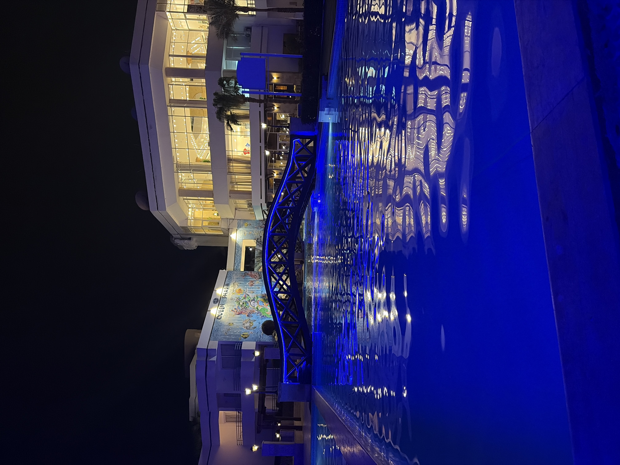 The image depicts a nighttime scene at the Queen Sharm Resort in Sharm El Sheikh, Egypt. A blue-lit pedestrian bridge spans over a body of water, possibly a pool or a decorative water feature. The water reflects the lights from the resort and the bridge.
The resort building is visible behind the bridge, with multiple floors and windows illuminated. A mural with marine life imagery is visible on the side of the building. Palm trees are located near the upper floor of the building. The foreground shows the water’s edge, and the scene is predominantly lit by blue lighting.