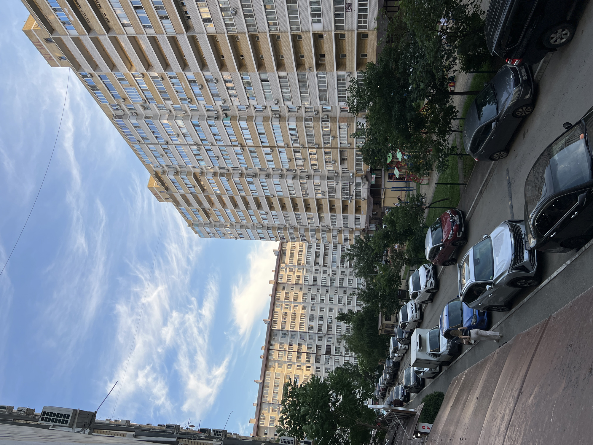 The image depicts a street scene in Krasnodar, Russia, featuring a row of parked cars and two apartment buildings. The perspective is from a slightly elevated position, looking down at the street and up at the buildings.
The dominant subjects are the cars, parked along the side of the street. Several are visible, including dark SUVs, a silver sedan, a blue hatchback, and a red car. The scene also includes mature trees lining the street, casting shadows on the pavement. The apartment buildings are tall and multi-story, characteristic of urban residential architecture. One building is lighter in color with balconies and numerous windows, while the other appears to be a more uniform grey concrete structure.
The sky is a clear blue with wispy white clouds, suggesting it is daytime, likely late afternoon or early evening given the light. There is no obvious activity or event taking place; the cars are parked, and the buildings appear to be residential.
Notable details include the presence of air conditioning units mounted on the balconies of the apartment buildings. Some of the cars have license plates visible, though the text is not clearly legible. A person is visible near one of the cars, appearing to be walking or interacting with the vehicle. The overall impression is of a typical urban residential area on a pleasant day.