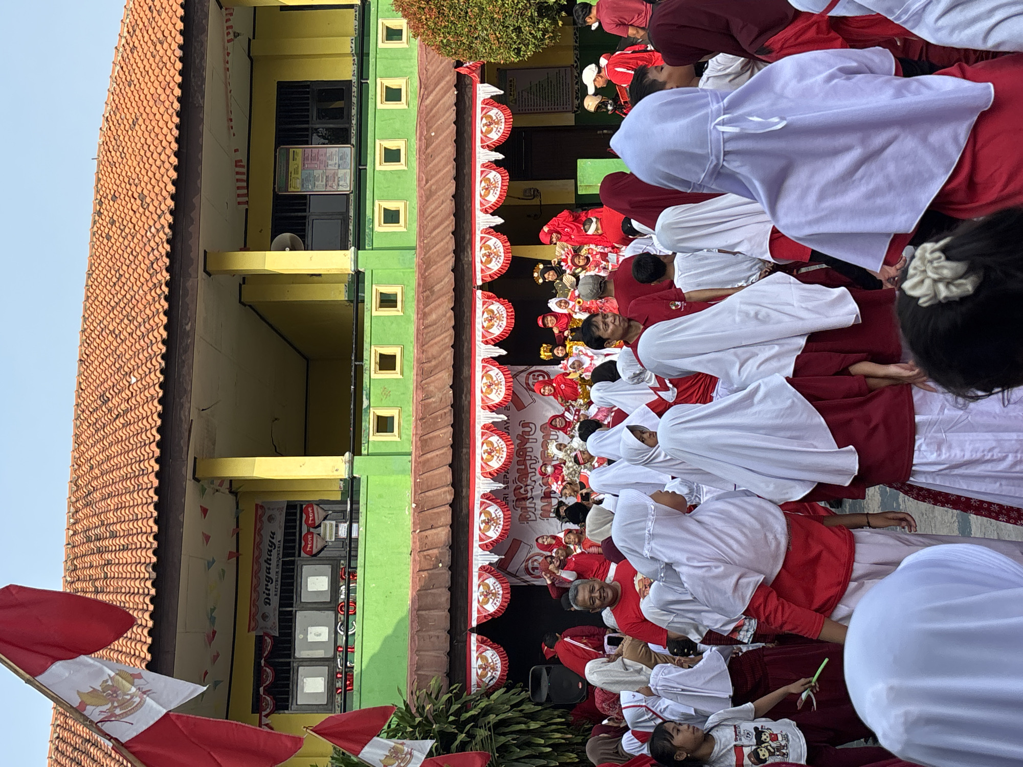 An outdoor event is taking place in front of a building that appears to be a school. The building has a yellow upper story and a green lower story with several small square windows. The roof is tiled in an orange-brown color. Red and white flags, featuring the Indonesian Garuda Pancasila emblem, are displayed prominently, including a large one on a flagpole to the left of the image and smaller ones decorating the facade of the building. A banner with text, possibly congratulating Indonesia, is visible above a stage area.
A crowd of people, many of whom are wearing red and white attire, are gathered. Several individuals, particularly the children and some women, are wearing white headscarves (hijabs). Some of the children appear to be participating in a performance on the stage, dressed in traditional costumes. The atmosphere suggests a celebration, likely related to Indonesian Independence Day given the national colors and celebratory banner. The presence of speakers on the left indicates a public address or performance. The foreground shows a dense crowd of onlookers facing the stage. The scene is brightly lit by sunlight, casting some shadows. The ground appears to be paved or a hard surface.