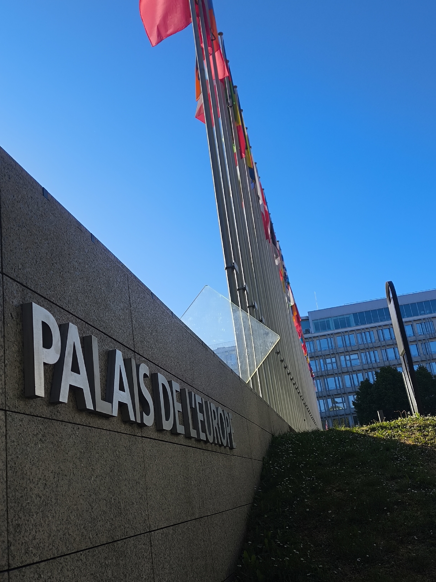 The image captures an upward-angled view of the exterior of the Palais de l'Europe in Strasbourg, France, under a brilliant, cloudless blue sky. In the foreground, the lower section of a robust, grey stone wall dominates the left, featuring large, metallic, three-dimensional letters that spell out "PALAIS DE L'EUROPE". The bright sunlight illuminates the letters and the upper edge of the wall, creating sharp contrasts and shadows.
Extending from the middle-ground towards the right and upwards is a long array of tall, silver flagpoles, adorned with numerous colorful flags, several of which are a vivid red, fluttering against the clear sky. These flags suggest the international character of the building. Behind the flagpoles, a modern, multi-storied building with a grid-like pattern of blue-tinted windows is partially visible. A dark, slender pole stands on the far right. Below the flagpoles and to the right, a verdant grassy slope, speckled with small white flowers, rises towards the building.
The bright natural light and the absence of clouds indicate a sunny day, likely morning or afternoon. The scene conveys a sense of institutional importance and international presence, fitting for the Council of Europe's headquarters in Strasbourg.
