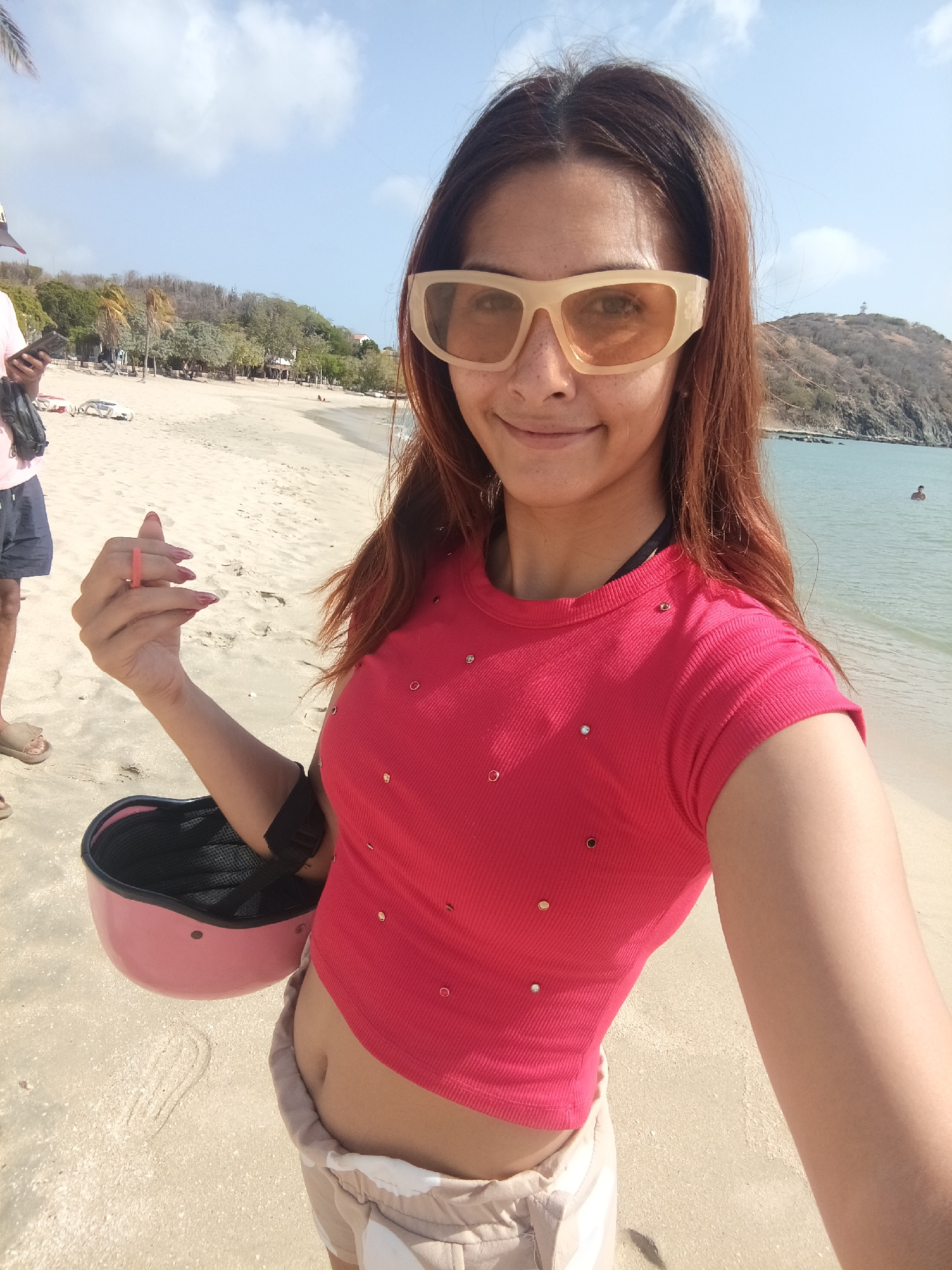 A fair-skinned woman is holding a pink helmet on a sandy beach with an ocean backdrop in Pedro González, Venezuela. The woman has long, reddish-brown hair, wearing rectangular light-colored sunglasses and a short-sleeved pink top with small embellishments, revealing part of her midriff. She is also wearing beige shorts. She is holding out one hand showing long red manicured nails. To her left, a person wearing a light-colored shirt and dark shorts is partially visible. The background includes a beach with palm trees and structures, a body of water, and a distant hill topped with a small structure. The sky is light blue with scattered clouds.