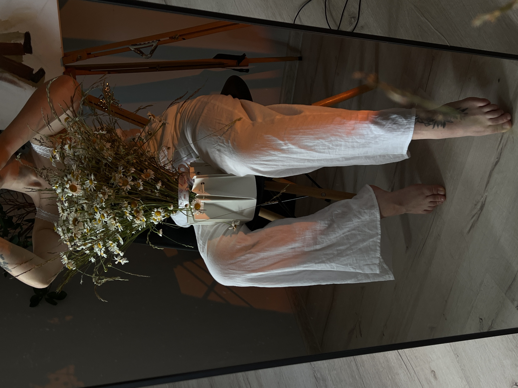 The image is a mirror reflection of a person sitting on a chair, holding a bouquet of daisies and greenery. The person is wearing white linen pants and a white top. The left leg of the person is visible from the knee down, with a tattoo on the foot. The person's right leg is mostly obscured by the white pants. A white vase holds the bouquet. The background is dark, with shadows and a wooden floor. An orange light casts a glow on the person's leg and the surrounding area. The scene appears to be indoors. This image is not specific to Ternopil, Ukraine.