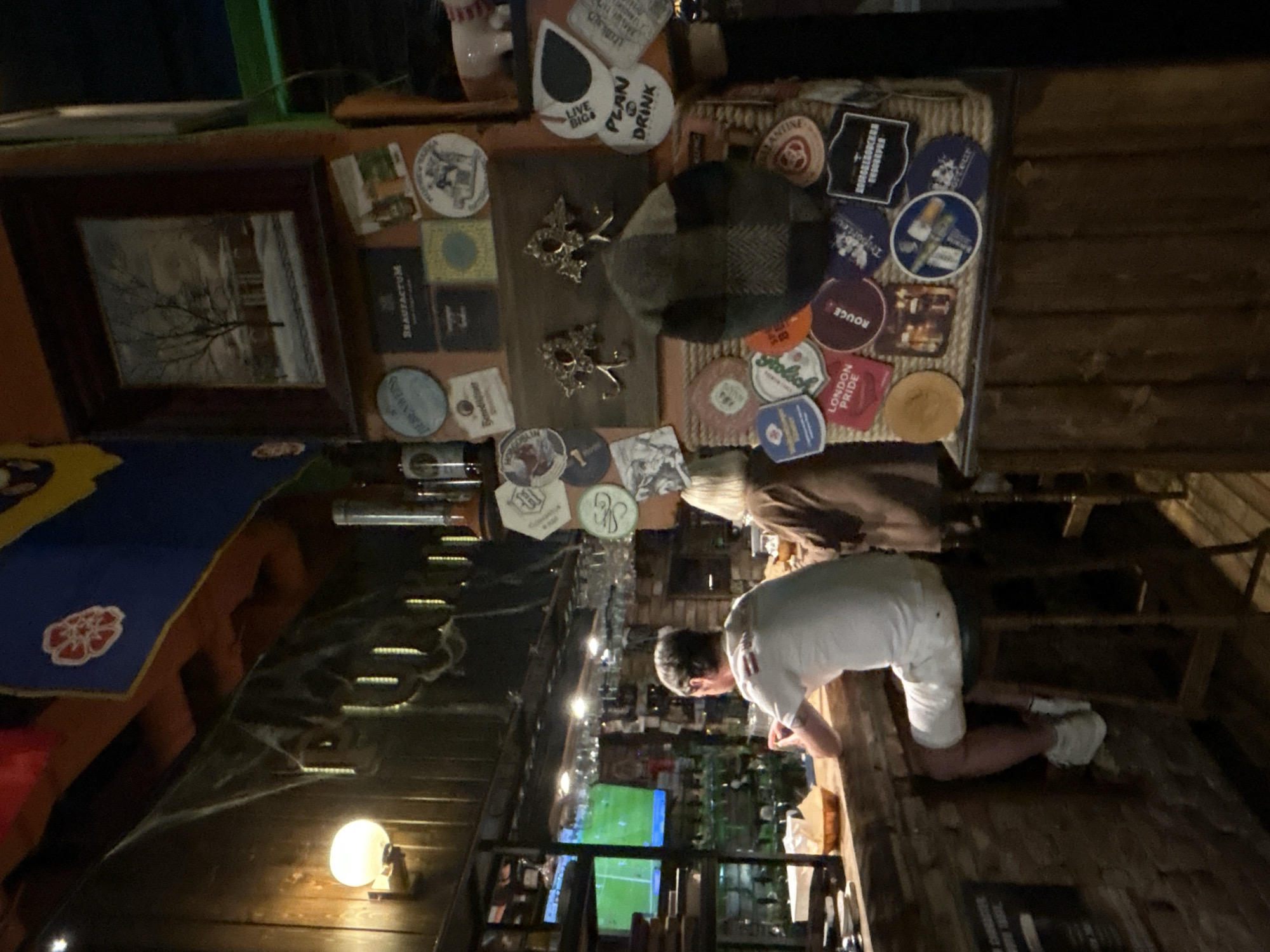 The image depicts the interior of a dimly lit, rustic bar. In the foreground, a man in a white t-shirt and white shorts sits on a stool at the bar, leaning on his forearms. To his right, a woman with blonde hair, wearing a brown jacket, sits at the bar facing away from the camera. The bar itself is made of brick and wood, with various bottles and glasses visible behind it, as well as a television screen showing a sports game. The walls are decorated with numerous beer coasters, framed pictures, and what appear to be hanging spiderweb decorations, suggesting a Halloween theme or general gothic aesthetic. The overall ambiance is that of a casual, possibly European-style pub. No specific details pinpoint the location to Moscow, Russia.