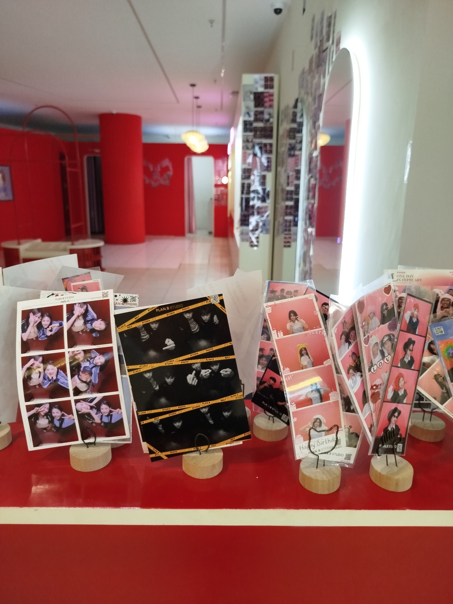 Captured in Da Lat, Vietnam, the photograph displays a vibrant red interior space adorned with photo booth strips displayed on wooden stands. The strips feature various subjects, including groups of friends and individuals, and are branded with "PLAN B STUDIO." The red color palette dominates the room, seen on the columns, walls, and counters, creating a visually arresting environment. An illuminated, arched doorway is visible in the background, alongside other decorative elements and a long hallway leading deeper into the building. The layout suggests the location is a photo studio or entertainment venue with a focus on self-photography and personalized photo strips.