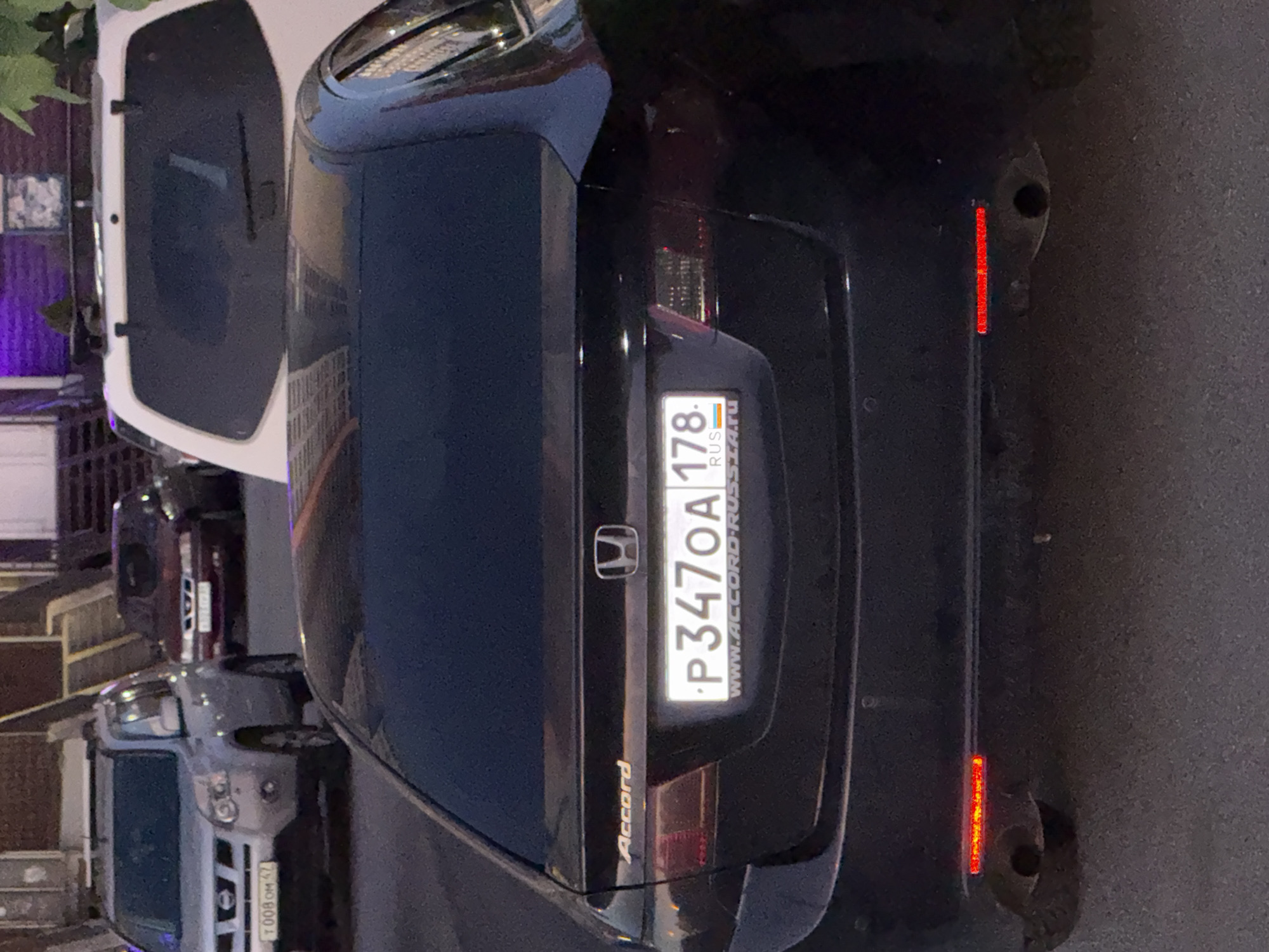 The image captures the rear view of a dark-colored Honda Accord sedan parked on a paved surface, likely a street or parking lot. The car's license plate is clearly visible and reads "P347OA 178". Below the license plate, a website address "WWW.ACCORD-RUSS.RU" is also displayed. The car has tinted windows and red reflectors on its bumper. To the left of the Accord, a silver Nissan X-Trail SUV is parked, with its license plate showing "T008OM 47". Behind the Nissan, a dark red sedan is partially visible. In the background, a white SUV is parked, and further back, a building with purple lighting is seen. The scene appears to be in an urban environment, possibly a residential area or a commercial district, characteristic of Saint Petersburg, Russia, given the license plate format. There are no people or animals visible in the frame. The lighting suggests it is either late afternoon or early evening.
