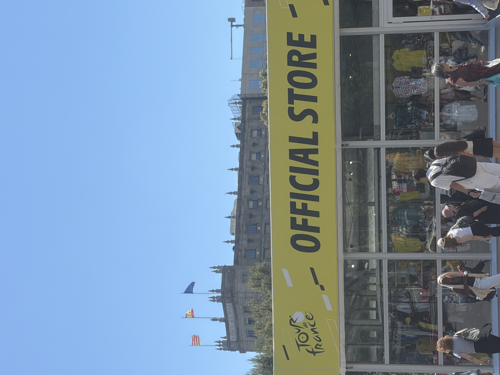 The image shows a street-level view of the "Tour de France" official store in Barcelona, Spain. The store has a large, vertical yellow sign with black lettering. The front of the store is lined with glass windows, displaying merchandise like jerseys.
Outside the store, several pedestrians are present, wearing various styles of clothing. They appear to be browsing or passing by.
In the background, across the street, the "Banco de España" building is visible, with flags atop its facade. The sky is a clear blue. Additionally, there's a building with a tower next to the "Banco de España".