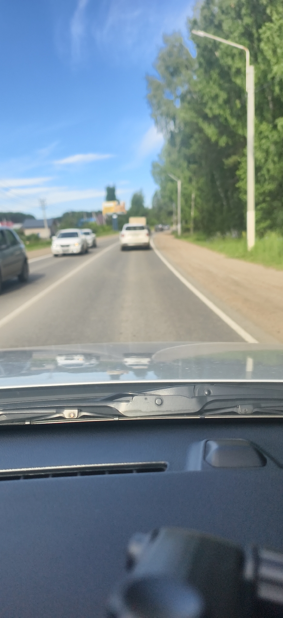 The image is a point-of-view shot from inside a vehicle looking through the windshield. The vehicle is on a paved road with a white dividing line. Several white cars are visible on the road ahead, some in motion. To the right of the road, there is a strip of grass and then a dense line of green trees. A tall, white street lamp is prominent on the right side of the road. In the distance, on the left side of the road, there are buildings and what appears to be a yellow structure with a steeple, possibly a church or a tall tower. The sky is blue with scattered white clouds. The foreground shows a portion of the vehicle's dashboard and windshield wipers. The overall scene suggests a drive on a sunny day in a semi-urban or rural area. Given the context of Chaikovsky, Russia, this could be a view on a road leading into or out of the city, possibly along a highway or main road surrounded by nature.