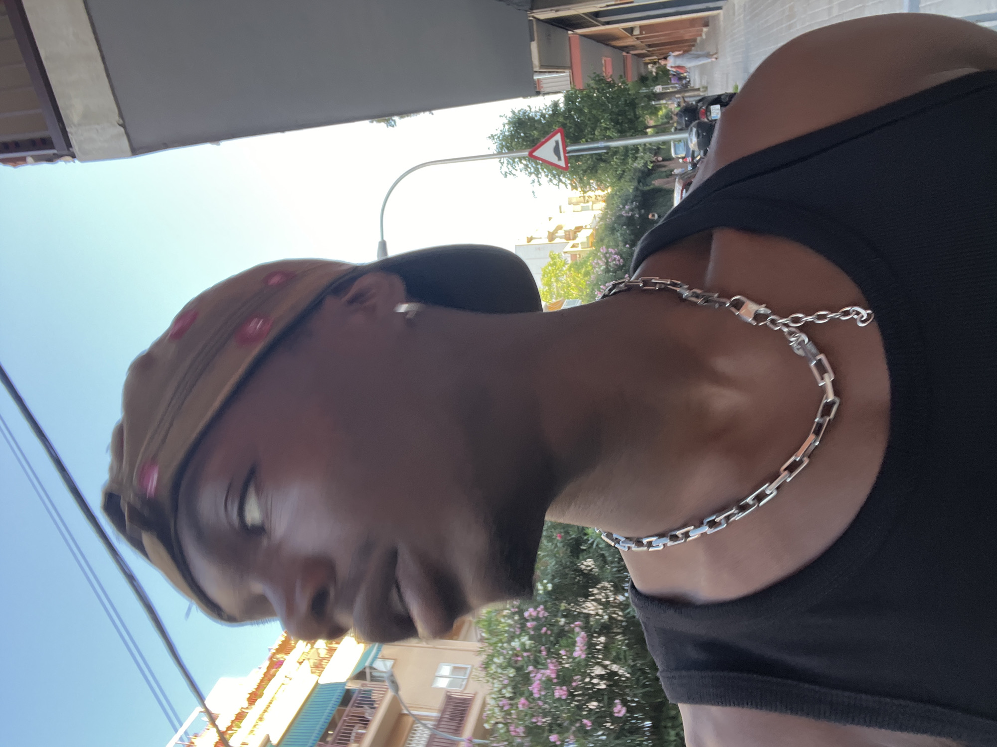 The image is oriented 90 degrees clockwise, presenting a profile view of a person outdoors under a clear, bright sky. The subject, a person with dark skin, is positioned on the left side of the frame, looking slightly downwards. They are wearing a light brown cap with several small, round, reddish-pink details on it, and a black, ribbed tank top. A prominent silver chain necklace with rectangular links and a visible clasp adorns their neck. A small earring is also visible.
The background reveals an urban setting, consistent with Barcelona, Spain. Above the person's head, the bright blue sky is visible, with parts of what appears to be an awning or building structure in the top left corner. Behind the person, light-colored buildings with balconies and some having a teal-colored facade or awning are discernible. Lush green trees and bushes with clusters of pink flowers, possibly bougainvillea or oleander, are scattered throughout the scene. A red-bordered, white triangular yield sign, characteristic of European road signs, is clearly visible on a pole in the mid-ground. Faint outlines of parked motorcycles or scooters and distant figures suggest a lively street or public area. The bright daylight and clear sky indicate a sunny day, likely midday or afternoon. No specific text is legible beyond the symbol on the yield sign.