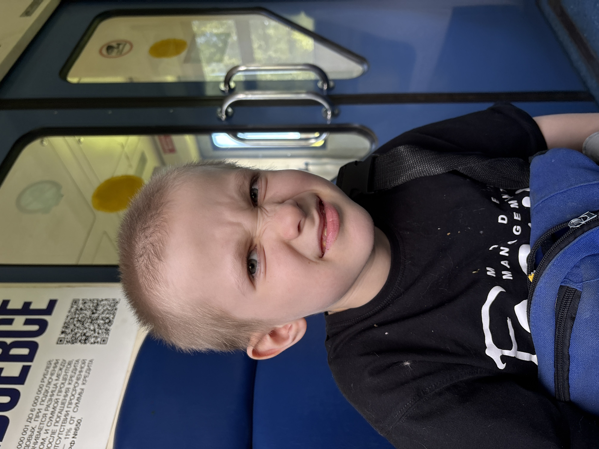 A medium close-up shot captures a young Caucasian boy, approximately 5-8 years old, inside what appears to be a public transport vehicle located in Красная Поляна, Russia. The boy is centered in the frame, making a distinctive, scrunched facial expression, characterized by a wrinkled nose, narrowed light-colored eyes, and slightly parted lips revealing some teeth. His short, light-blonde hair is visible. He is dressed in a black short-sleeved t-shirt featuring a partially visible white graphic print on the chest, which includes the fragmented words "F M MAN G". A black strap, consistent with a backpack or shoulder bag, crosses his right shoulder, leading to a blue bag resting on his lap, secured by a visible black zipper.
The background to the boy's left consists of a royal blue upholstered seat or wall. Above his head, a white sign with black Russian text and a QR code is partially visible. The text includes financial figures and terms, notably "000 001 ДО 6 000 000 РУБЛЕЙ" and the large word "ВСЁ" (VSE) at the uppermost edge. To the boy's right, two large, vertical windows with dark blue frames are visible, suggesting a door or partition. Two silver, curved grab handles are prominently affixed to the right window panel. Through the windows, blurred green foliage and bright sunlight indicate an outdoor environment, suggesting the vehicle is either moving or parked outdoors. Yellow circular stickers are present on the windowpanes.