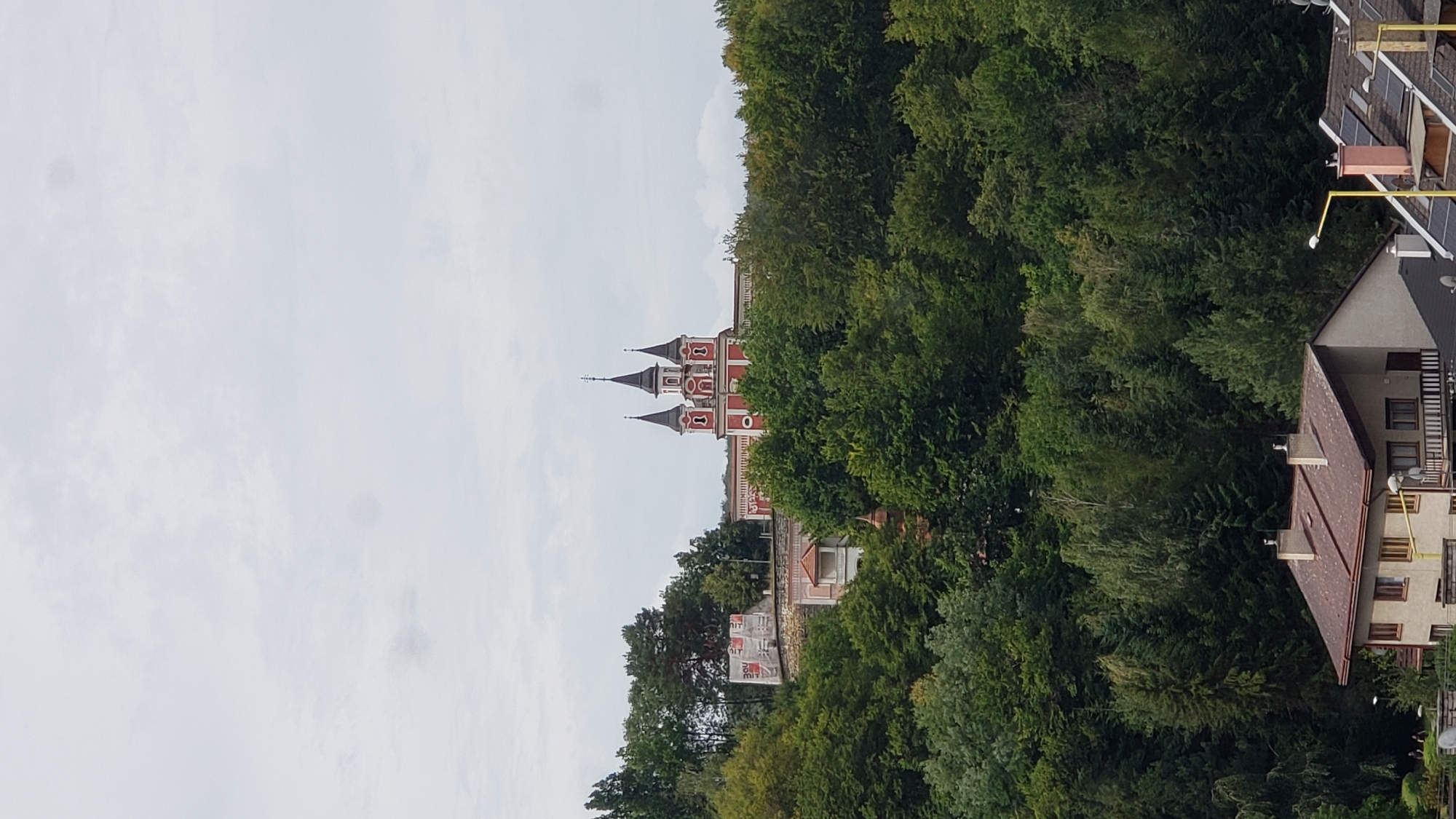 The image captures a view of the Church of Saint Nicholas (Kostol svätého Mikuláša) in Prešov, Slovakia, perched on a hill amidst lush green trees. The church is a striking red building with multiple spires, characteristic of its architectural style. Below the church, the dense foliage of a forest covers the hillside. In the foreground, a residential building with a reddish-brown roof is partially visible, suggesting the church is situated in an area with both natural landscapes and urban development. The sky is overcast with a light gray hue.