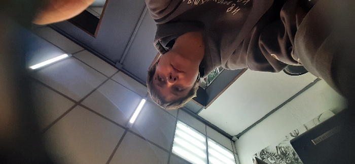 An indoor, low-angle photograph depicts a young individual and their immediate surroundings. The specific location within the city of Saratov, Russia, cannot be determined from the visual evidence.
**People:** One individual, appearing young, with short, dark brown hair, is positioned in the center-left foreground, looking upwards and to the right. The person wears a grey hooded sweatshirt with light-colored text partially visible on the chest. A small metallic piercing is present in the left nostril, and a small earring is visible in the left earlobe. The person's right arm, covered by the sweatshirt sleeve, is partially visible in the bottom right corner of the frame.
**Subjects/Objects:** In the upper left background, a dark object, possibly a laptop or a binder, is visible, partially obscuring a light-colored wall. Above this, a black and white drawing or print of a stylized face or head is affixed to the wall. The wall beneath this print features a dark rectangular object with light text, partially legible as "TOLOGI." To the left and behind the individual, a dark, vertical shelving unit or cabinet is discernible.
**Environment:** The setting is an indoor space with a light-colored suspended ceiling composed of square panels. Several illuminated fluorescent strip lights are mounted on the ceiling. The walls vary in color; the wall to the left is light-colored, while the wall to the right appears darker, possibly grey or dark blue, with vertical structural elements or paneling. A dark framed opening or doorway is visible in the far right background.
**Actions/Interactions:** The individual is posing for a photograph, likely a selfie, given the low camera angle and the partial view of their arm. No other individuals or interactions are depicted.
