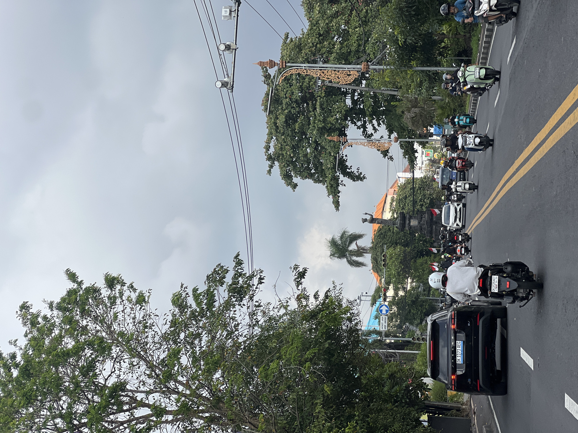 This is a photograph taken from a vehicle on a street in Denpasar, Indonesia, likely during the daytime given the lighting conditions, though the sky is overcast with heavy clouds. The street is paved and has yellow double lines marking the lanes. A black SUV, possibly a Range Rover, is in the left lane. Several motorcycles and scooters are on the road. One scooter rider in the foreground wears a white helmet and a light-colored shirt. In the background, there are trees lining the street, a statue or monument, buildings, and overhead power lines with streetlights. Traffic lights are visible, one set showing red. A street sign with Indonesian text and an arrow pointing right is also present. The overall scene depicts a busy urban street with motorized traffic.