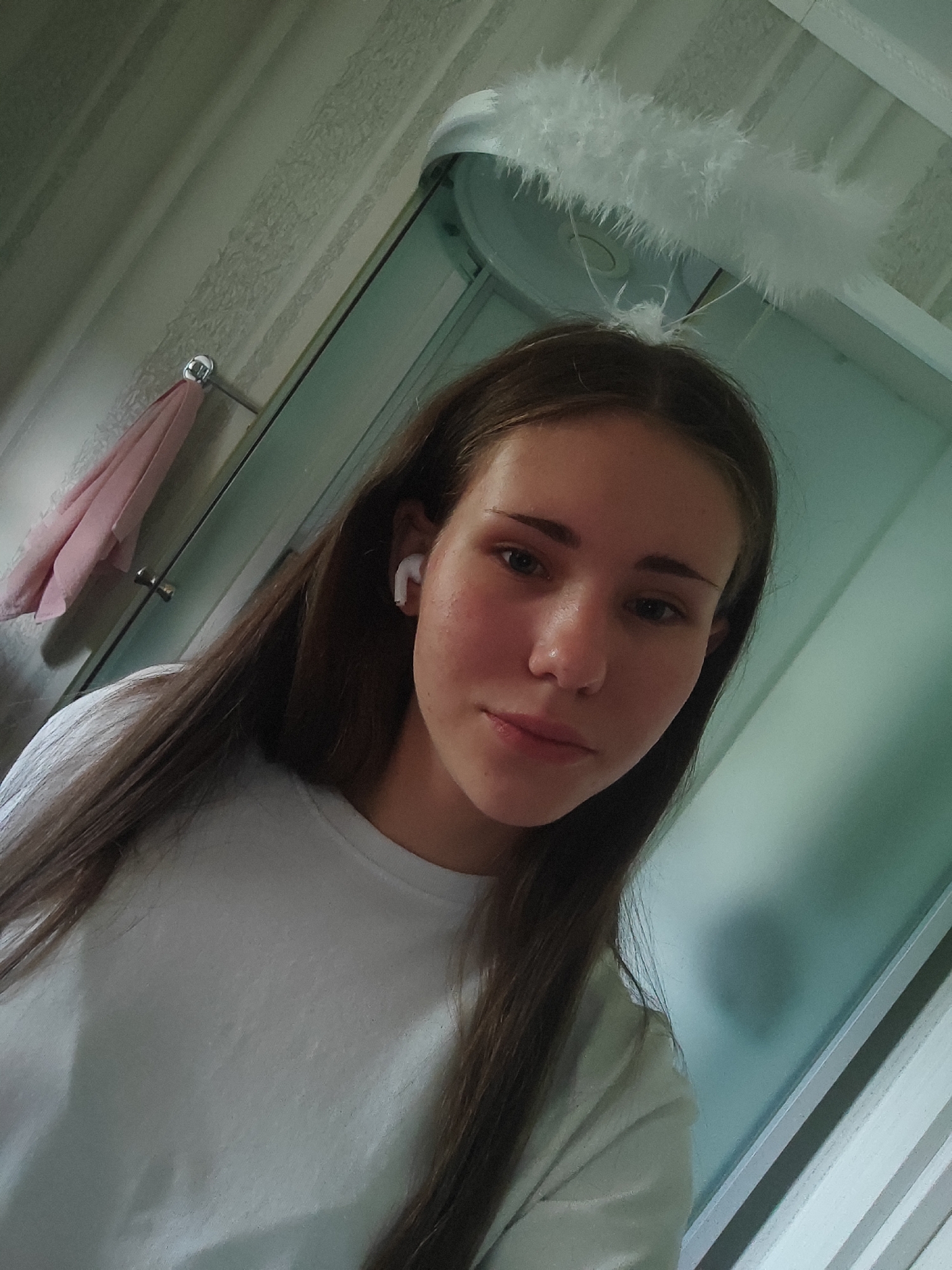 The image shows a young woman indoors, likely in a bathroom, wearing a white t-shirt and white wireless earbuds. She has long brown hair and is wearing a fluffy white headband that resembles an angel's halo. The background includes a tiled wall and a glass shower door. The lighting suggests it is daytime. No specific location in Svietlahorsk, Belarus can be determined from the image.