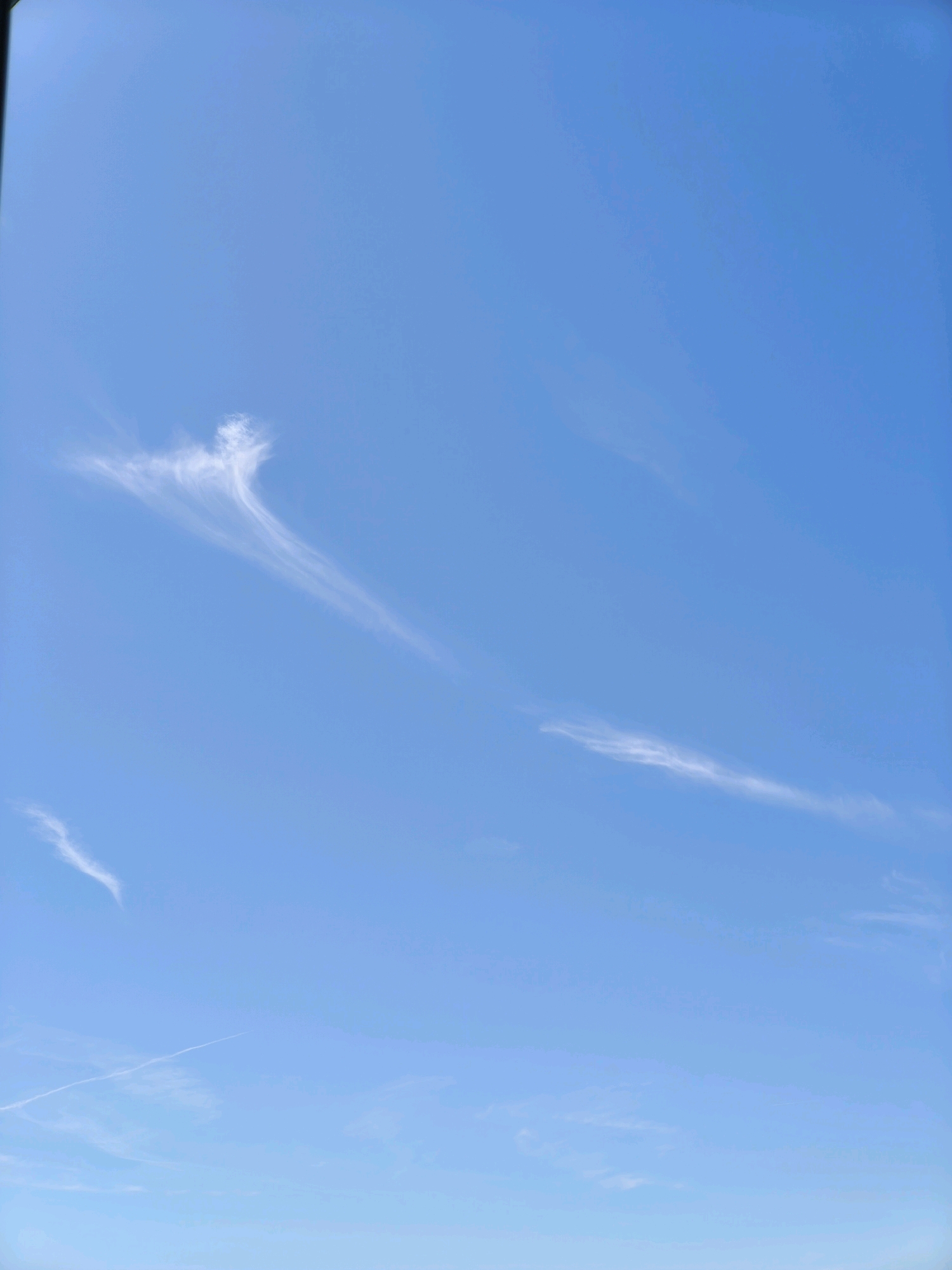 A clear blue sky with wispy white clouds is visible. One cloud formation, located in the upper left quadrant of the image, resembles an angel with outstretched arms and a flowing robe. Another thin, elongated cloud stretches diagonally across the mid-right portion of the sky. A faint contrail is visible in the lower left corner, indicating the passage of an aircraft. The image appears to be taken from Passignano sul Trasimeno, Italy, on a bright, clear day.