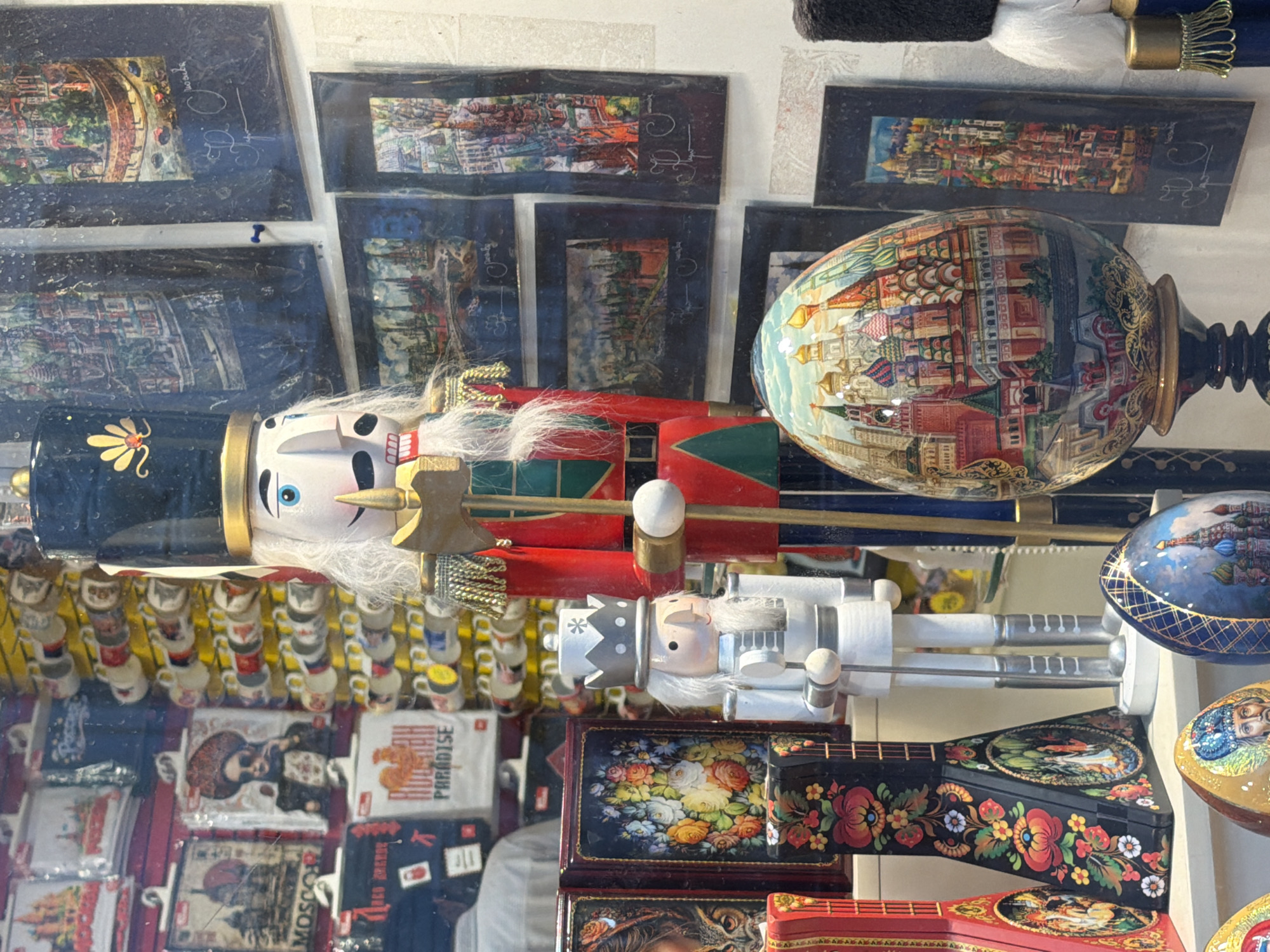 The image displays a collection of Russian-themed souvenirs, likely from a market or shop. In the foreground, a large wooden nutcracker with a black hat and red uniform is prominently featured. To its right, another nutcracker, this one adorned with a crown and white uniform, is visible. These figures appear to be decorative rather than in use.
Behind the nutcrackers, the scene is filled with various items. A large, intricately painted egg, possibly a Faberge-style egg, showcases a detailed depiction of a cityscape, likely St. Basil's Cathedral. The egg is placed on a dark stand with ornate golden detailing. Below the egg and nutcrackers, several smaller decorative items are arranged, including what appear to be miniature cups stacked on yellow stands and several flat, rectangular items with colorful painted scenes, possibly lacquer boxes or trays. Some of these depict floral arrangements and other traditional Russian motifs.
Further into the background, there are framed prints or paintings hanging on a wall, also featuring cityscapes and traditional artwork. The overall impression is one of a vibrant display of cultural artifacts. The lighting suggests an indoor setting, possibly with artificial light or ambient daylight filtering through a window, as there are reflections on some of the surfaces. No specific time of day or weather conditions can be determined.
Visible text includes "MOSCOW" on one of the flat items. Another item has the word "PARADISE" printed on it. There are also some handwritten characters on some of the framed prints.