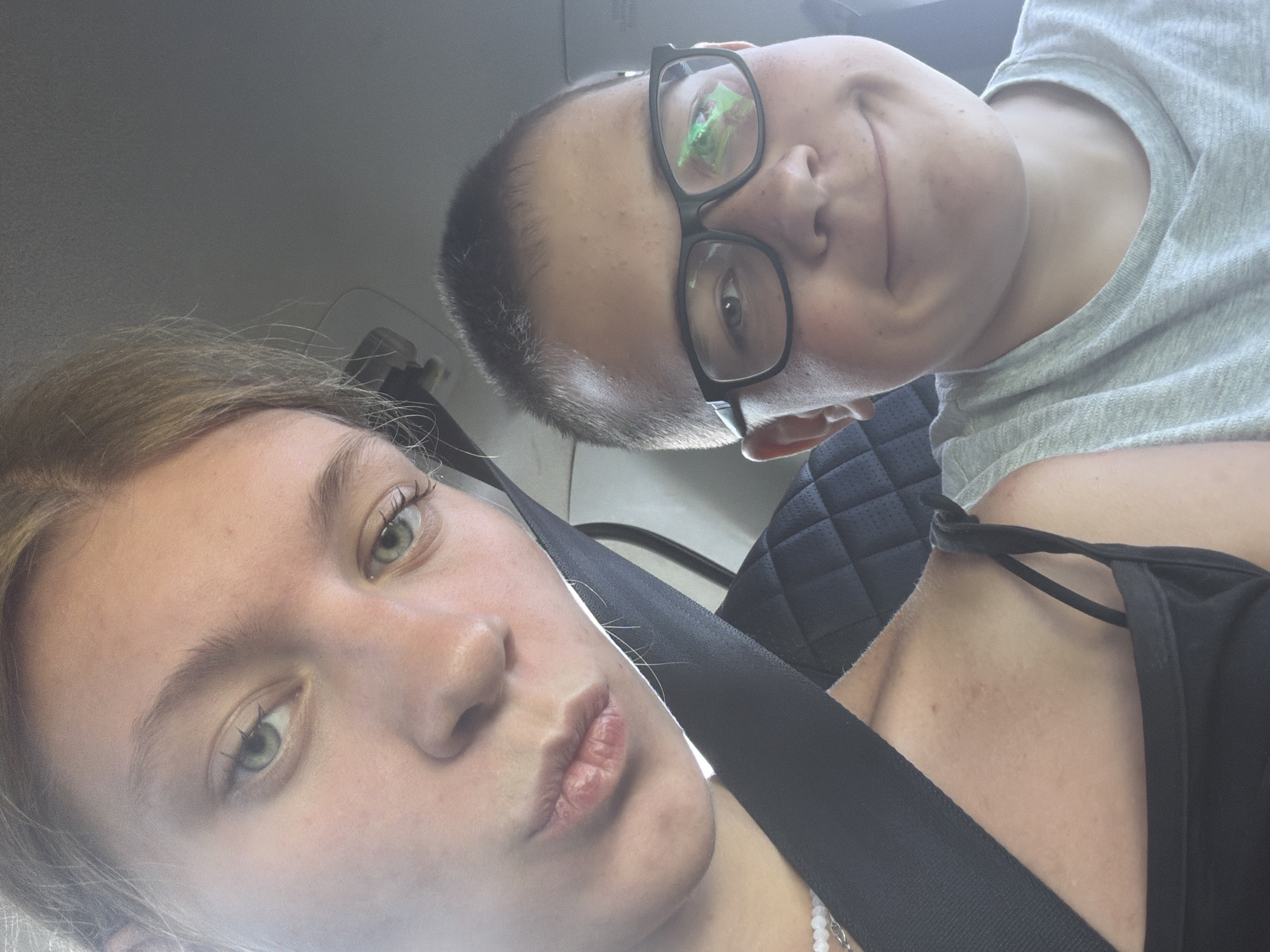 A selfie taken inside a vehicle shows two individuals. On the left, a young female with blonde hair pulled back is in the foreground, angled towards the camera with her lips pursed. She is wearing a black top and a seatbelt. To her right and slightly behind her, a young male with short brown hair and glasses is also looking at the camera and smiling. He is wearing a light gray t-shirt. The background appears to be the interior of a car, with a seatbelt buckle visible on the right. The image quality is slightly blurred, suggesting it was taken quickly. The scene is consistent with being in a vehicle, potentially traveling. The location in Novoe Feizullovo, Russia, is not directly indicated by any visual cues in the image.