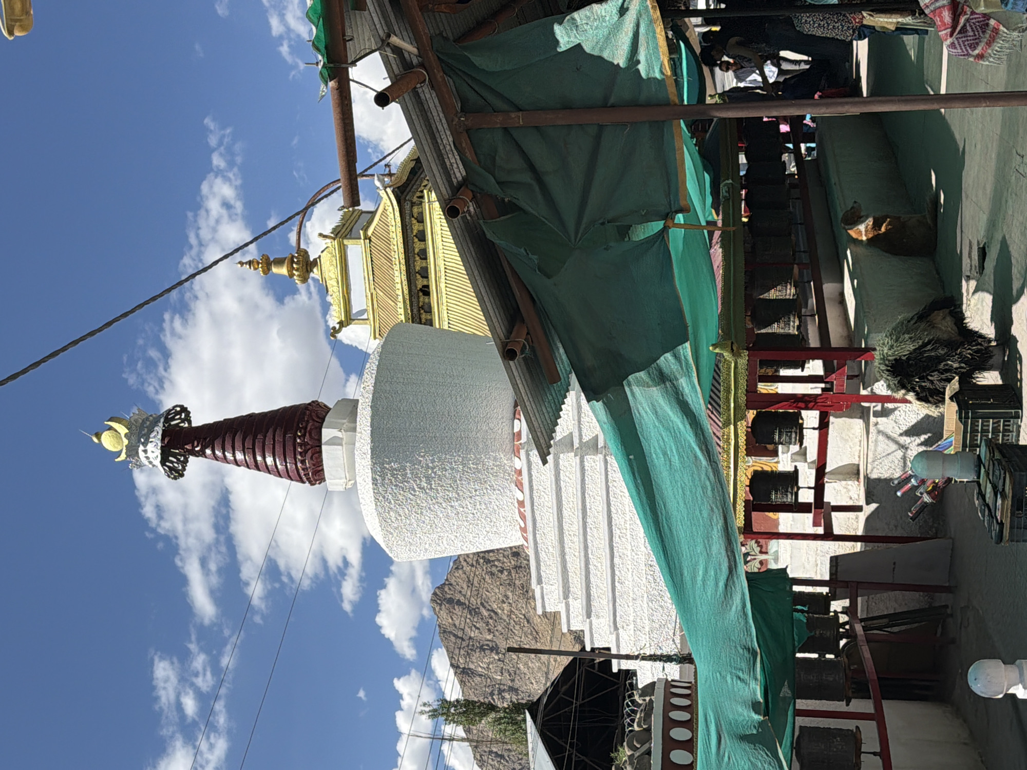 The image depicts a Buddhist stupa in Leh, India, under a bright, partly cloudy sky. The stupa is predominantly white with red and gold accents on its spire. To the right of the stupa, a golden-roofed building with intricate detailing is visible. In the foreground, a makeshift shelter covered with green tarpaulin offers shade. This area is equipped with rows of prayer wheels mounted on a platform. A dog is seen sitting against a wall, and several people are present, some appearing to be tourists. The environment suggests a place of religious and cultural significance, likely a monastery or temple complex in Leh. The presence of prayer wheels and the architectural style of the stupa and temple are characteristic of Buddhist sites in the region.