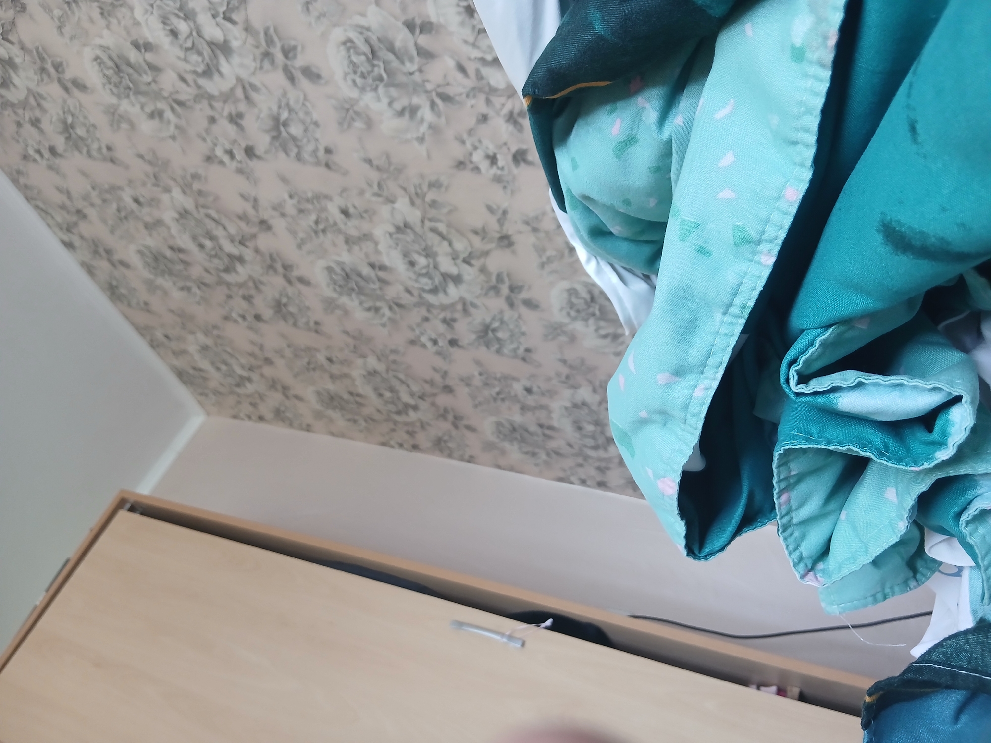 The image shows an interior scene with a floral-patterned wallpaper on the ceiling. A light-colored wooden cabinet is visible in the lower portion of the frame. Draped over the cabinet is a pile of fabric in varying shades of green and blue, some with a patterned design. A thin, dark cable is also visible. The location is not discernible from the image. The city of Tsarevets, Bulgaria, cannot be confirmed as the location.