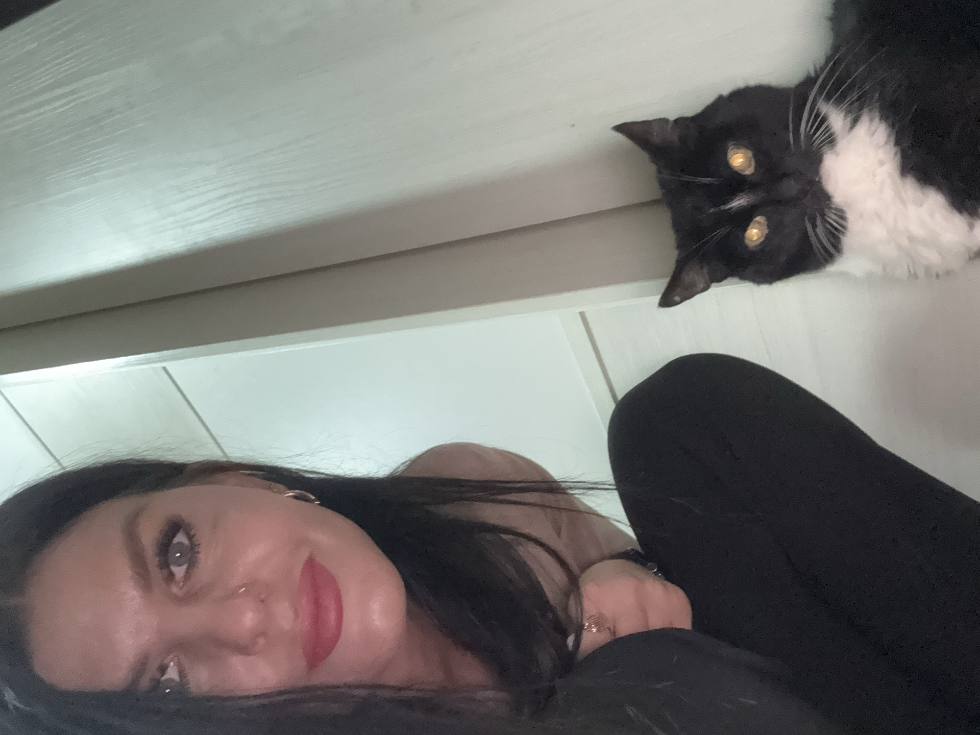 This image captures a low-angle perspective of a woman and a cat in an indoor setting. The composition is rotated, with the woman appearing in the lower left portion and the cat in the upper right.
The woman has long dark hair and is looking directly at the camera with a relaxed or slightly smiling expression. She has noticeable eye makeup and wears bright red lipstick. A nose piercing is visible on her left nostril. She appears to be reclining or lying down, with her body clad in dark fabric, possibly black leggings or pants, in the lower right.
The cat is a black and white tuxedo cat, also looking directly at the camera. Its distinctive bright yellow eyes stand out against its dark fur. The cat is positioned above and to the right of the woman, seemingly resting on a light-colored surface, possibly a shelf or furniture.
The background consists of light-toned surfaces, suggesting an interior space. A textured, possibly wooden, light-colored surface is prominent in the upper part of the image, with a darker horizontal line beneath it. The overall lighting suggests an indoor environment. No text is visible in the image.