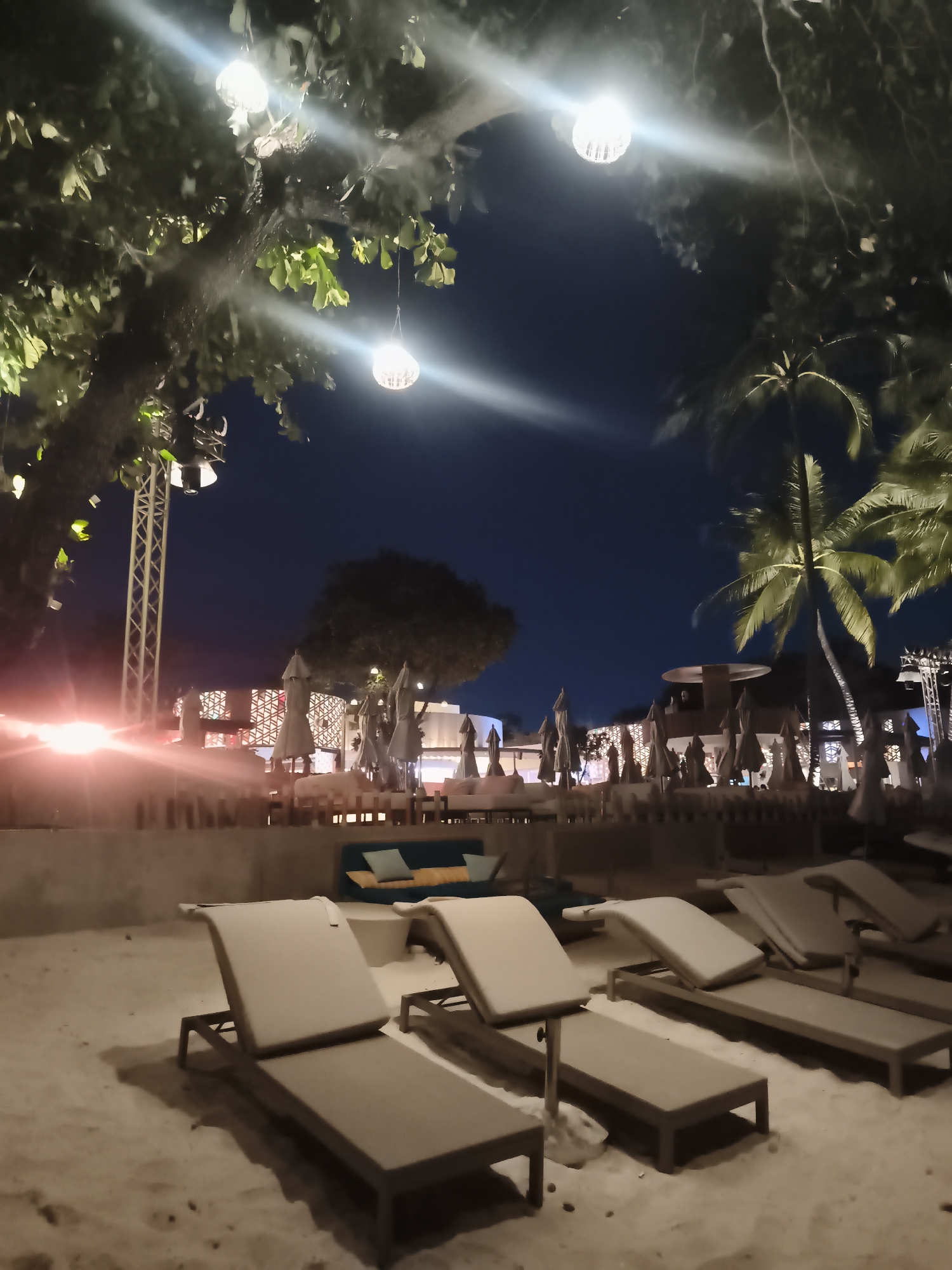 The image depicts a nighttime scene at a beach in Baan Chaweng, Thailand. In the foreground, several empty beach loungers are arranged on the sand. Behind the loungers, a low wall separates the beach from a slightly elevated area with sofas and closed beach umbrellas. Further back, there is what appears to be a building with a modern design, illuminated by soft lights. Palm trees are visible on the right side of the frame, their fronds illuminated by artificial light. Above, decorative light bulbs hang from tree branches, creating a warm glow. In the left of the image, there's a metal structure with lights attached. The sky is dark, suggesting it is late in the evening. There is no apparent human activity.