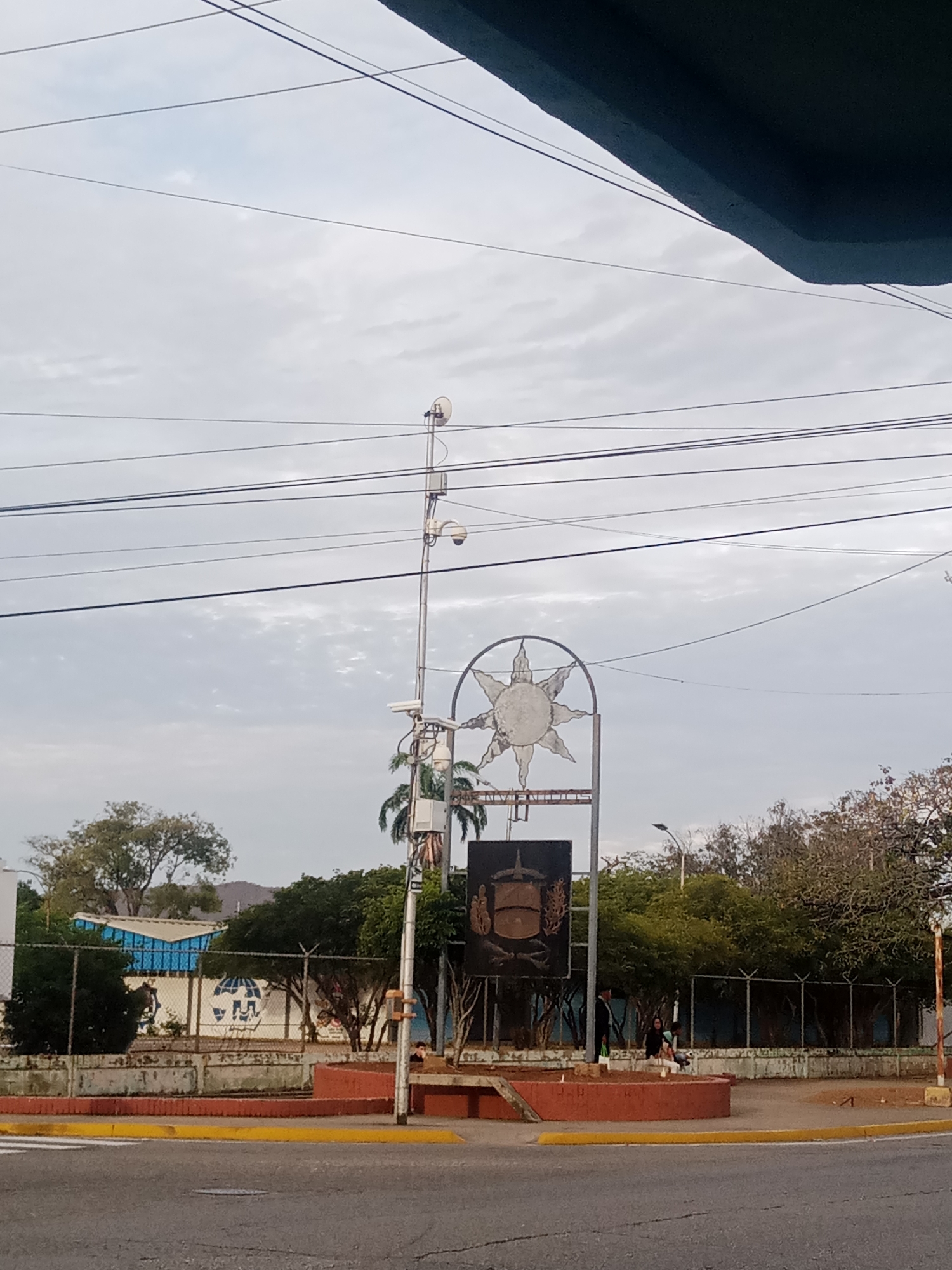 The image depicts a town square or plaza in Juan Griego, Venezuela. The scene is viewed from street level, showcasing a low angle toward the central monument.
A prominent metal structure with a stylized sun emblem is located at the heart of the plaza. A darker sign or plaque, presumably a memorial or marker, is situated beneath the sun emblem. A tall metal pole with multiple surveillance cameras is positioned nearby.
Surrounding the plaza is a low, red-painted concrete barrier with a yellow safety line. The perimeter of the plaza is lined with trees and shrubbery. A fence runs behind the trees.
A few people are present in the plaza. They are sitting near the marker.
Overhead, a network of electrical wires crisscrosses the sky. The sky is overcast.