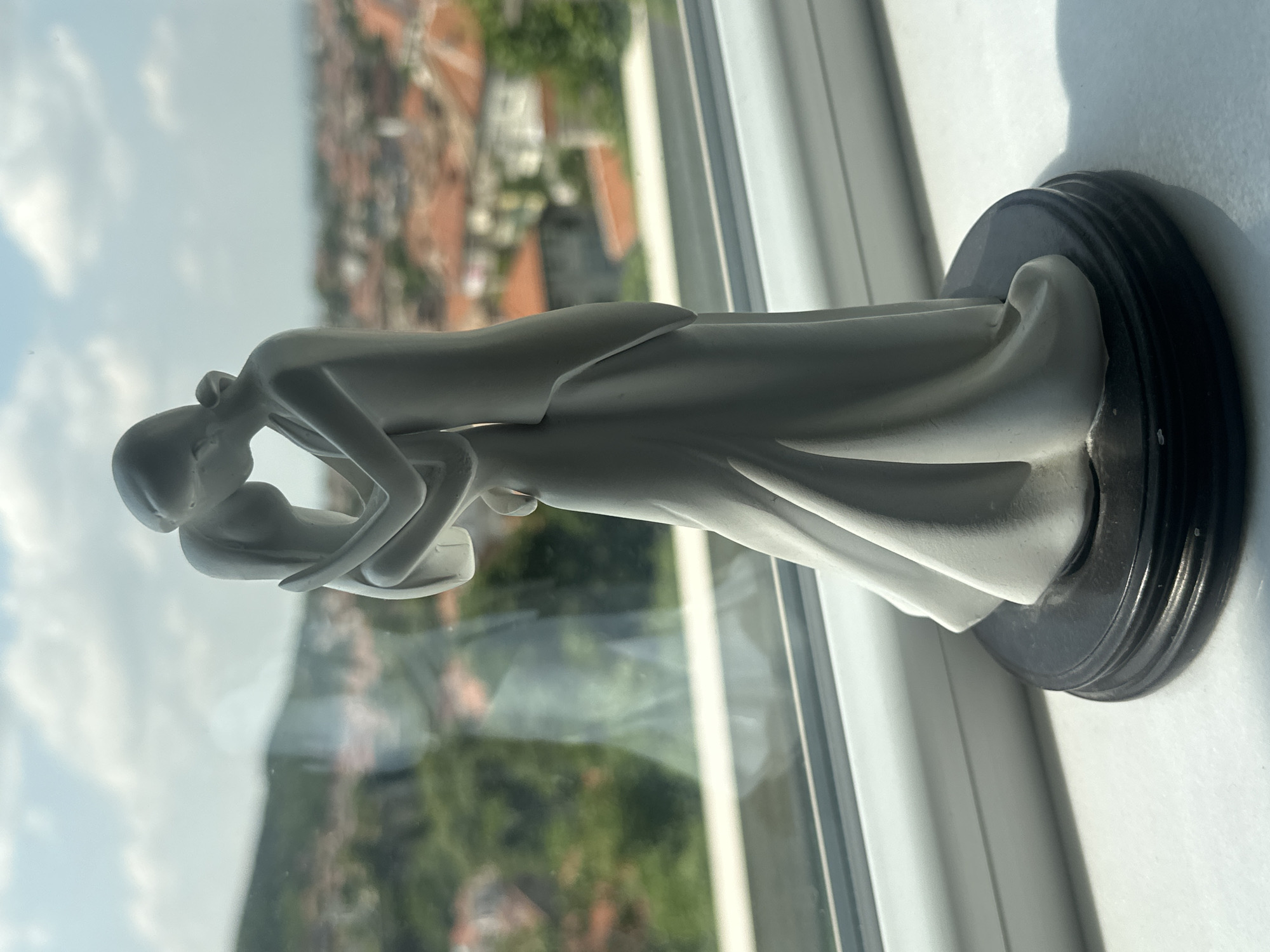 A light gray or white statue of a couple embracing and kissing is prominently displayed on a white windowsill, overlooking a townscape. The statue, depicting abstract figures of a man in a suit and a woman in a flowing dress, stands on a dark, circular, fluted base, conveying a sense of romance and affection.
Through the window, a clear daytime scene is visible, characterized by soft, natural light and partly cloudy skies. The background reveals architectural elements common in European towns, including buildings with reddish-brown roofs, interspersed with abundant green foliage and distant hills. This outdoor setting, viewed from what appears to be an interior window, places the scene in Asenovgrad, Bulgaria, as per the provided context. Shadows cast by the statue on the windowsill suggest the position of the sun. There is no visible text in the image.