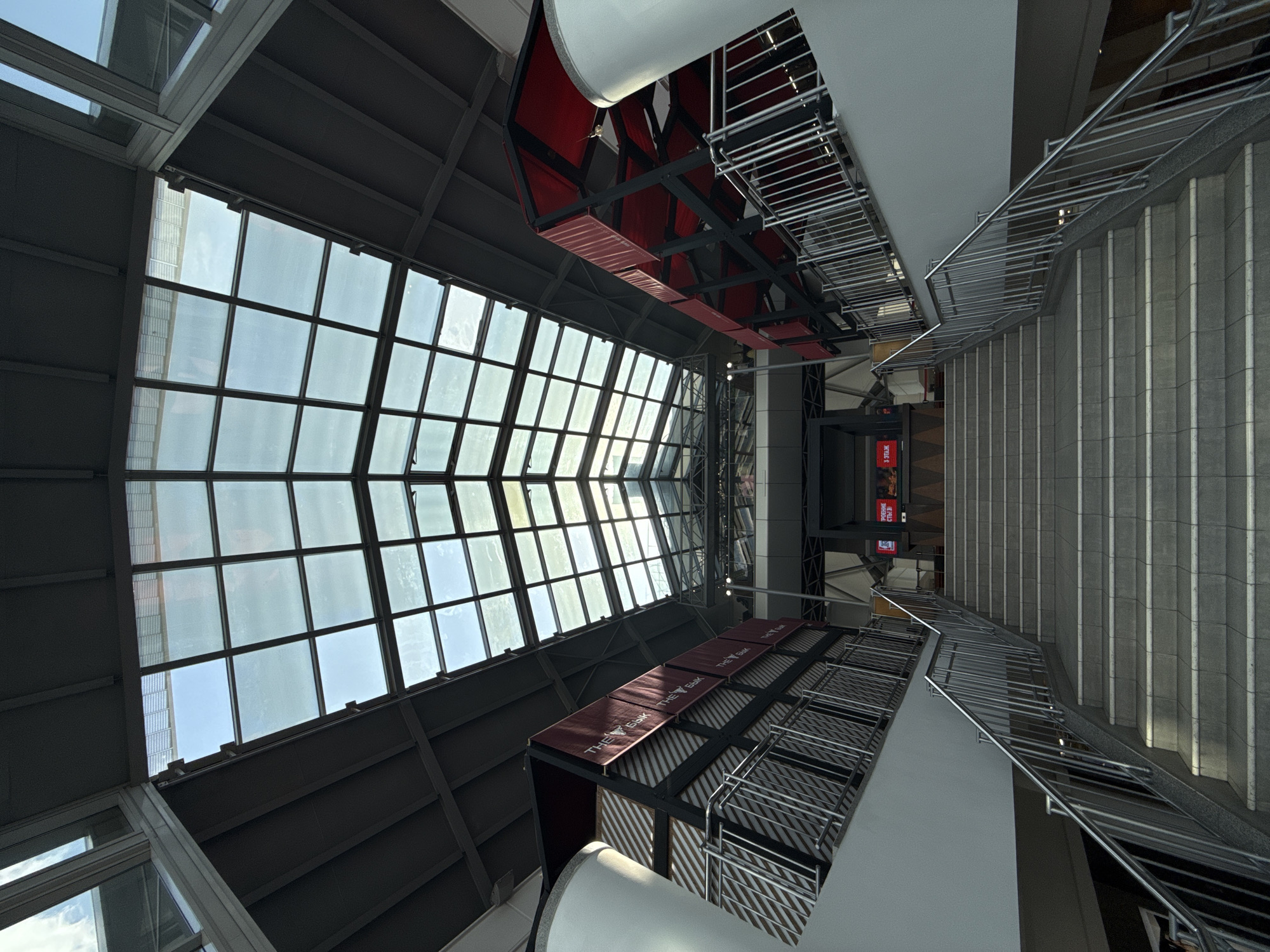 This image shows an interior view, looking up towards a large skylight in a building in Moscow, Russia. The skylight is composed of many square glass panes set in a grid-like frame. Below the skylight, there's an architectural structure with a maroon-colored covering featuring text "THE БЫК." The structure has a metal railing. To the right, there's a set of concrete stairs with a metal railing, ascending out of view. At the end of the stairs, a sign reading "ПОДЪЕМ СЛЕВА" is visible. The building has gray structural components.
