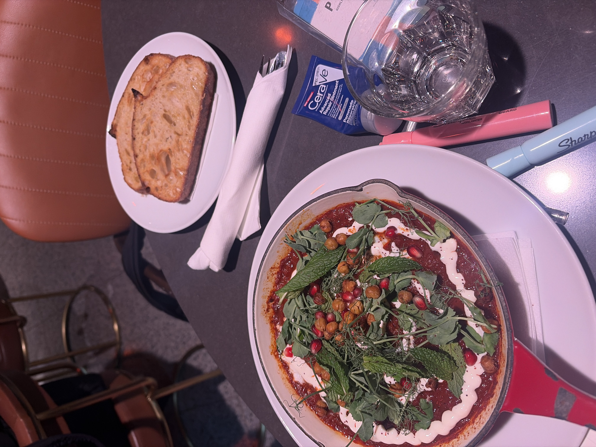 Here is a detailed description of the image:
**Setting:**
The image appears to be taken inside a restaurant or bar. Brown leather furniture, possibly seating, is visible.
**Objects:**
* **Food:** A white plate holds three slices of bread. A shallow skillet contains a dish with red sauce, chickpeas, pomegranate seeds, and a white creamy swirl, topped with green herbs (possibly mint).
* **Tableware:** A white folded napkin with cutlery. A glass of water.
* **Personal Items:** A tube of Cerave Advanced Repair Ointment, a pink highlighter, and a light blue Sharpie pen are lying on the table.
* A plastic wrapped item.
* The skillet is resting on a white plate with a red rim.
**Composition:**
The photograph is taken from a high angle, looking down at the table setting. The composition is somewhat cluttered with food, personal items, and tableware. The lighting is soft and possibly artificial.
**Overall Impression:**
The scene depicts a casual meal setting with personal items scattered on the table, suggesting a relaxed or busy atmosphere.