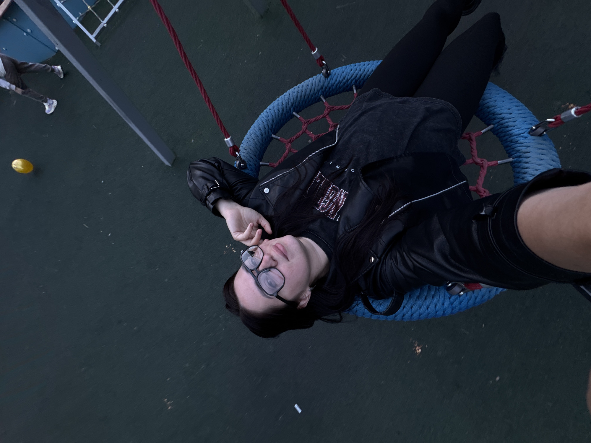 This high-angle, likely selfie, captures a person relaxing on a circular swing in an outdoor setting, possibly a playground. The main subject, a person with dark hair and glasses, is lying on their back on a large blue swing with red support ropes. They are wearing a black leather jacket, a dark t-shirt with partially visible red text on the front (appearing as "ON" or "OF NO"), and black leggings or tights. Their left arm is bent with the hand near their head, while their right arm extends out of frame, suggesting they are holding the camera.
The ground beneath is a dark, uniform surface, possibly rubberized flooring common in playgrounds. In the upper left background, the legs and lower torso of another person wearing dark pants and light-colored sneakers are visible, implying they are walking. A small yellow object, possibly a ball or toy, is on the ground to the left. Part of a metallic structure, consistent with playground equipment, is also visible in the background. A small white rectangular piece of litter is noticeable on the ground below the main subject's head.
The lighting appears soft and somewhat dim, suggesting either late afternoon, early evening, or an overcast day. The person on the swing conveys a sense of calm and relaxation. The scene depicts a moment of leisure in a public recreational area.
