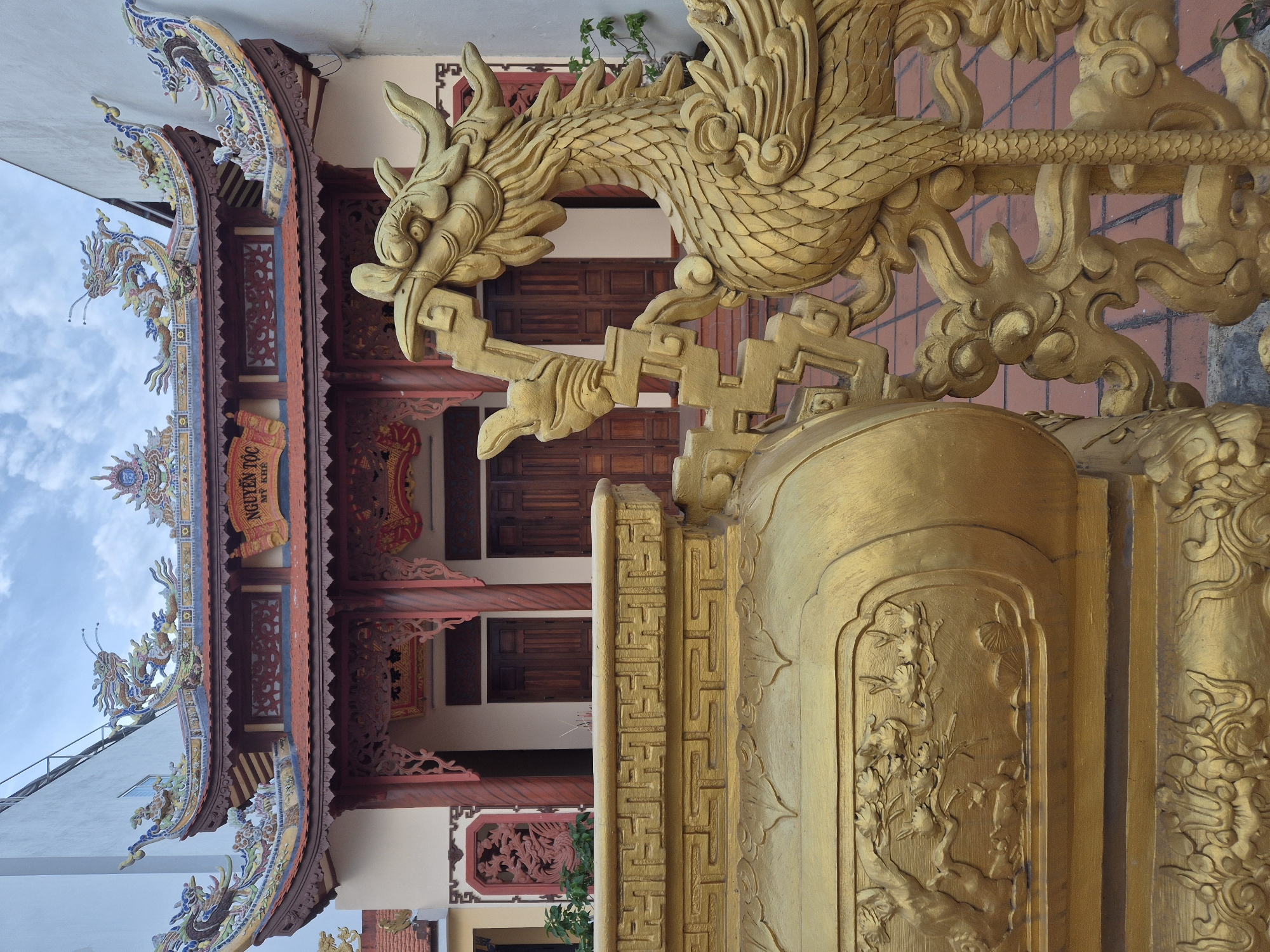 The image captures a close-up view of a traditional Vietnamese temple in the city of Đà Nẵng, Vietnam. In the foreground, a large, ornate golden urn is partially visible on the left, adorned with intricate carvings. To the right of the urn, a golden dragon statue stands guard, its head turned slightly towards the temple. Behind these golden artifacts, the entrance of the temple is visible, featuring double wooden doors and ornate architectural details. The temple roof is decorated with colorful ceramic dragons and intricate patterns, typical of Vietnamese temple architecture. The ground is paved with terracotta tiles. The background shows a cloudy sky and the white wall of an adjacent building. The scene appears peaceful and serene.