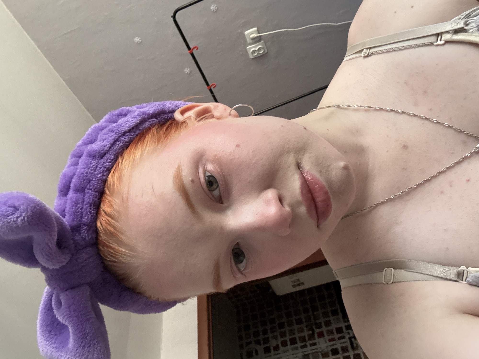 A close-up, tilted, indoor shot captures a fair-skinned person with red hair and a purple headband with bunny ears. They are wearing a thin silver necklace and a bra strap is visible on their shoulder. The background includes a white wall with an electrical outlet and a black metal frame.