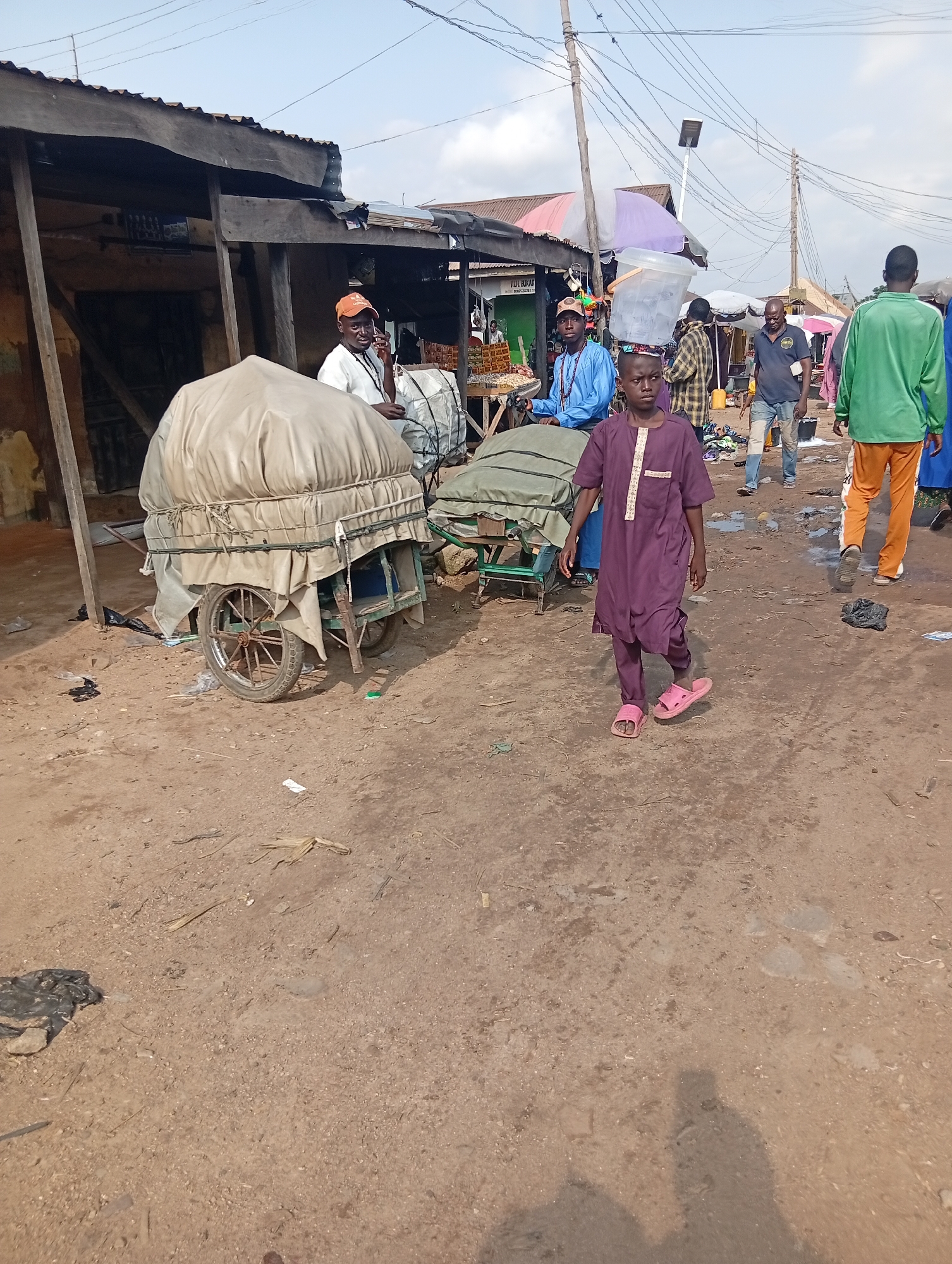 The image depicts a bustling outdoor market scene in Jalingo, Nigeria. In the foreground, a young boy in a purple traditional outfit walks towards the right, wearing pink sandals. Behind him and to the left, a man in an orange cap and white shirt is seen talking on a mobile phone, standing next to a large, covered cart with visible wheels. Further in the background, several other individuals are present, some appearing to be vendors. One man in a blue outfit is seated at a stall displaying goods. The environment is characterized by a dirt ground, with several stalls and structures made of wood and corrugated metal. Various carts and umbrellas are scattered throughout the market. The sky above is clear and bright, indicating daytime. The overall impression is one of a vibrant local marketplace.