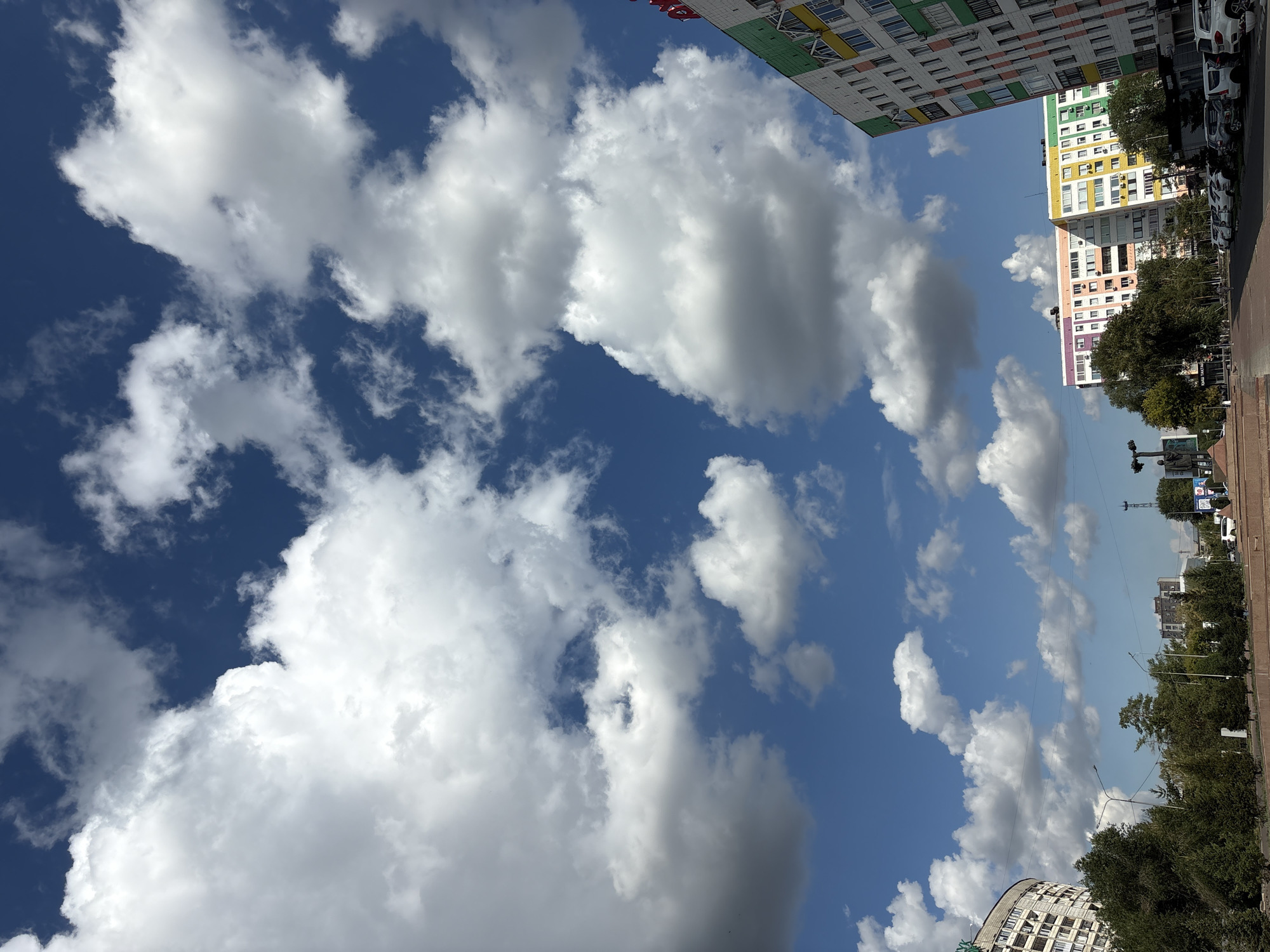 This is a street-level, eye-level, wide shot of a city street in Pavlodar, Kazakhstan, under a dynamic sky filled with large, white cumulus clouds against a deep blue background. The image is dominated by the sky, with buildings and trees occupying the lower third.
On the right side of the image, a tall, modern apartment building with a distinct green, white, and orange facade is prominently featured, angled towards the viewer. To the left of this building, other apartment buildings are visible, some with bright, multi-colored facades in shades of pink, orange, yellow, and green. These buildings appear to be residential blocks, typical of Soviet-era urban planning.
In the foreground, a paved street or sidewalk runs horizontally across the bottom. Several cars are parked along the right side of the street, mostly white SUVs and sedans. Trees with green foliage line the left side of the street, forming a green barrier between the sidewalk and the buildings further back. A statue of a man is visible on a pedestal on the left side of the frame, in front of a large, curved apartment building with many windows. Streetlights are also present along the sidewalk. The overall impression is of a residential and commercial area on a clear, cloudy day.
