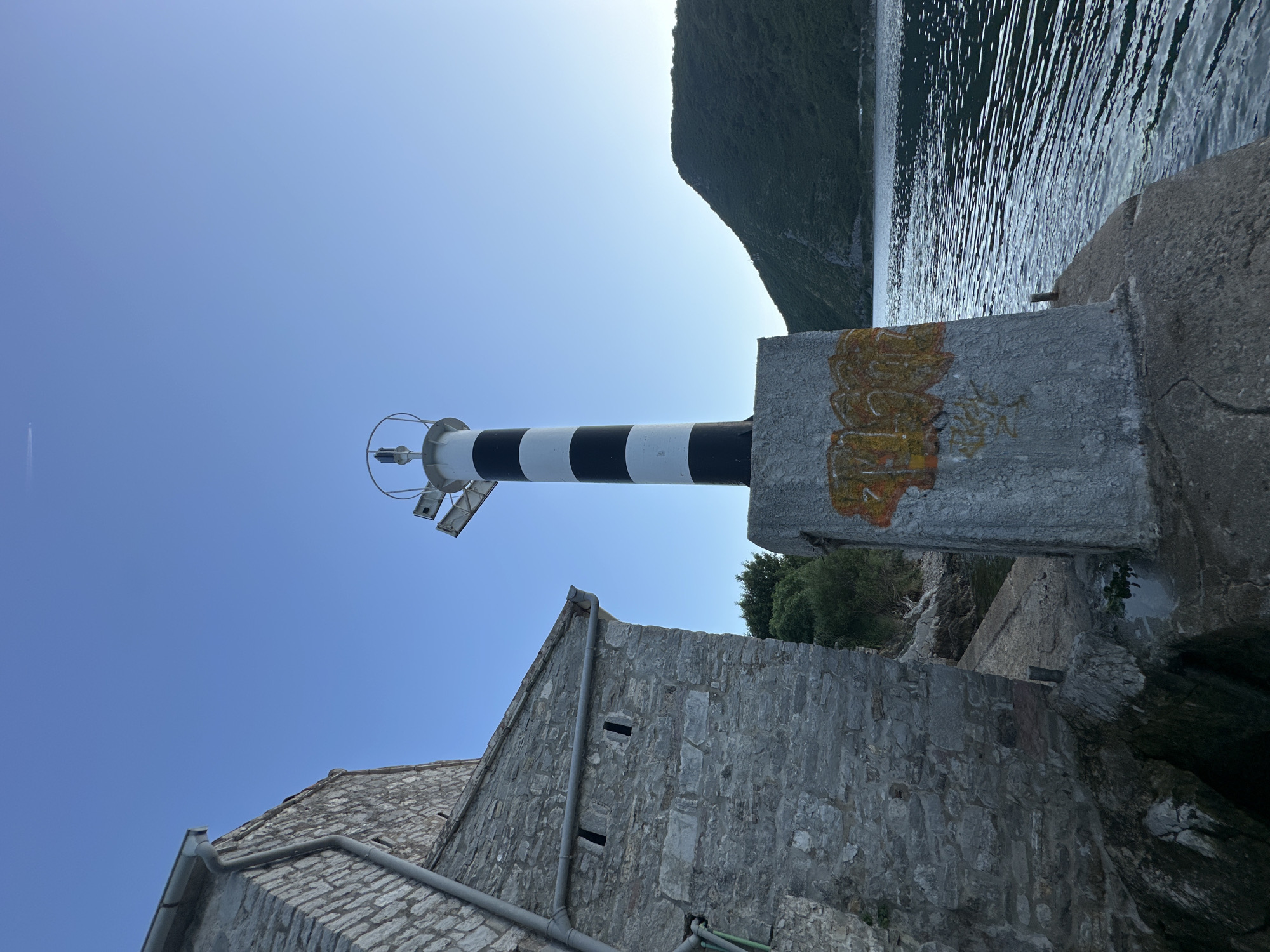 This image captures a coastal scene in Lepetane, Montenegro, featuring a black and white striped cylindrical lighthouse positioned on a concrete base to the right of an old stone building. The lighthouse's concrete base exhibits orange and yellow graffiti on its front surface. The top of the lighthouse features a circular metal railing and an apparatus that includes what appear to be solar panels. To the left, a multi-story building constructed from rough-hewn grey stone blocks features a metal gutter system running along its sloped roofline and down its side. Rectangular, narrow openings are visible on the stone wall of the building. The foreground comprises concrete and rocky terrain leading down to the water. The sea, a calm body of water with visible ripples, extends to the right and into the background. Across the water, a large, dark green, tree-covered mountain or hill dominates the horizon. The sky above is clear and light blue, with a faint white contrail from an aircraft visible in the upper left quadrant. No individuals are present in the scene.
