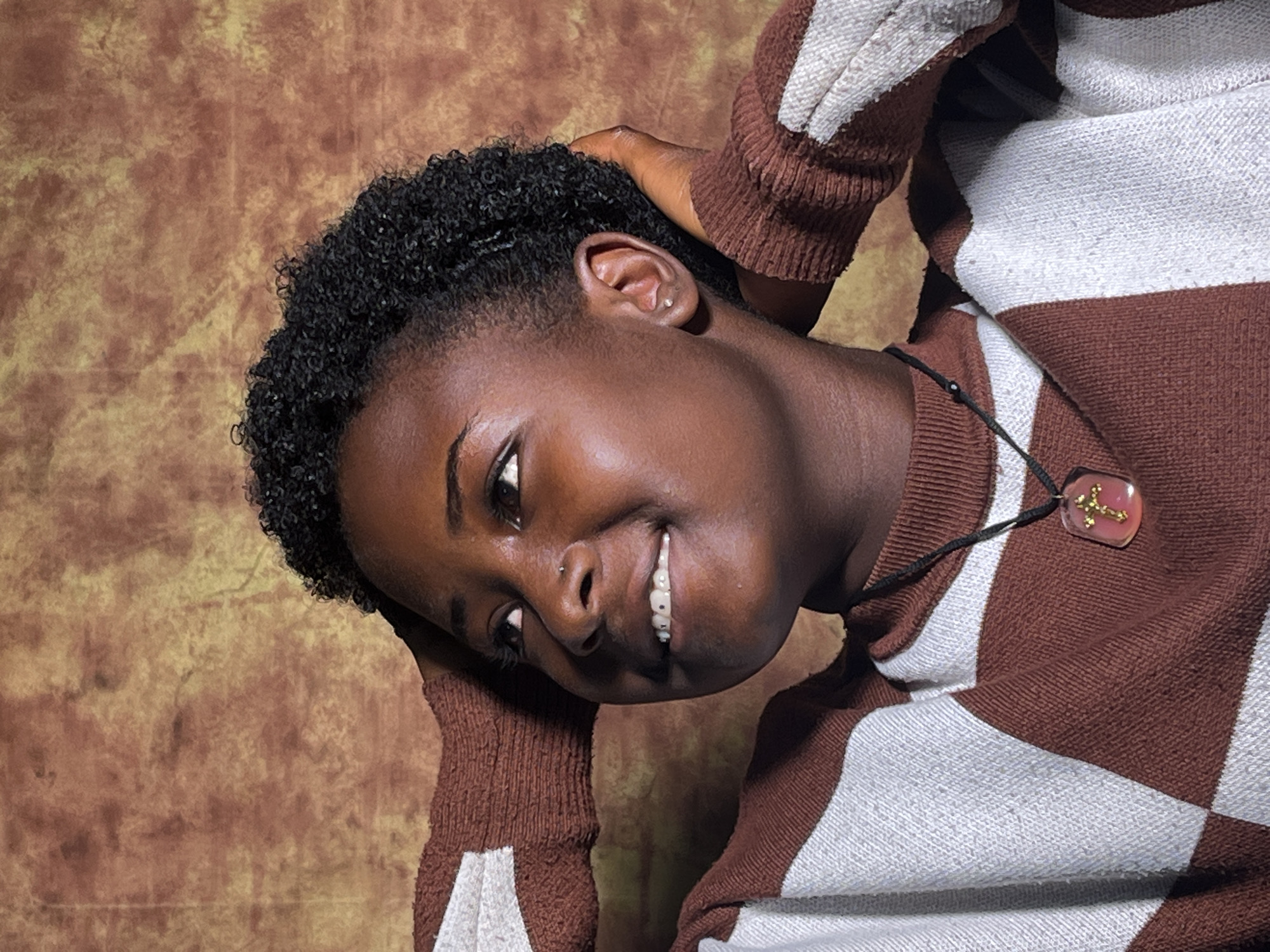A young Black person is shown from the chest up, looking upwards and to the left with a smile. They have short, curly black hair, a nose piercing, and braces visible on their teeth. Their hands are placed behind their head, resting on their hair. They are wearing a brown and white striped sweater and a necklace with a pink, translucent pendant containing a gold cross. The background is a mottled brown and gold textured wall. The image appears to be a studio portrait. The scene is located in Barakin Akawo, Nigeria.